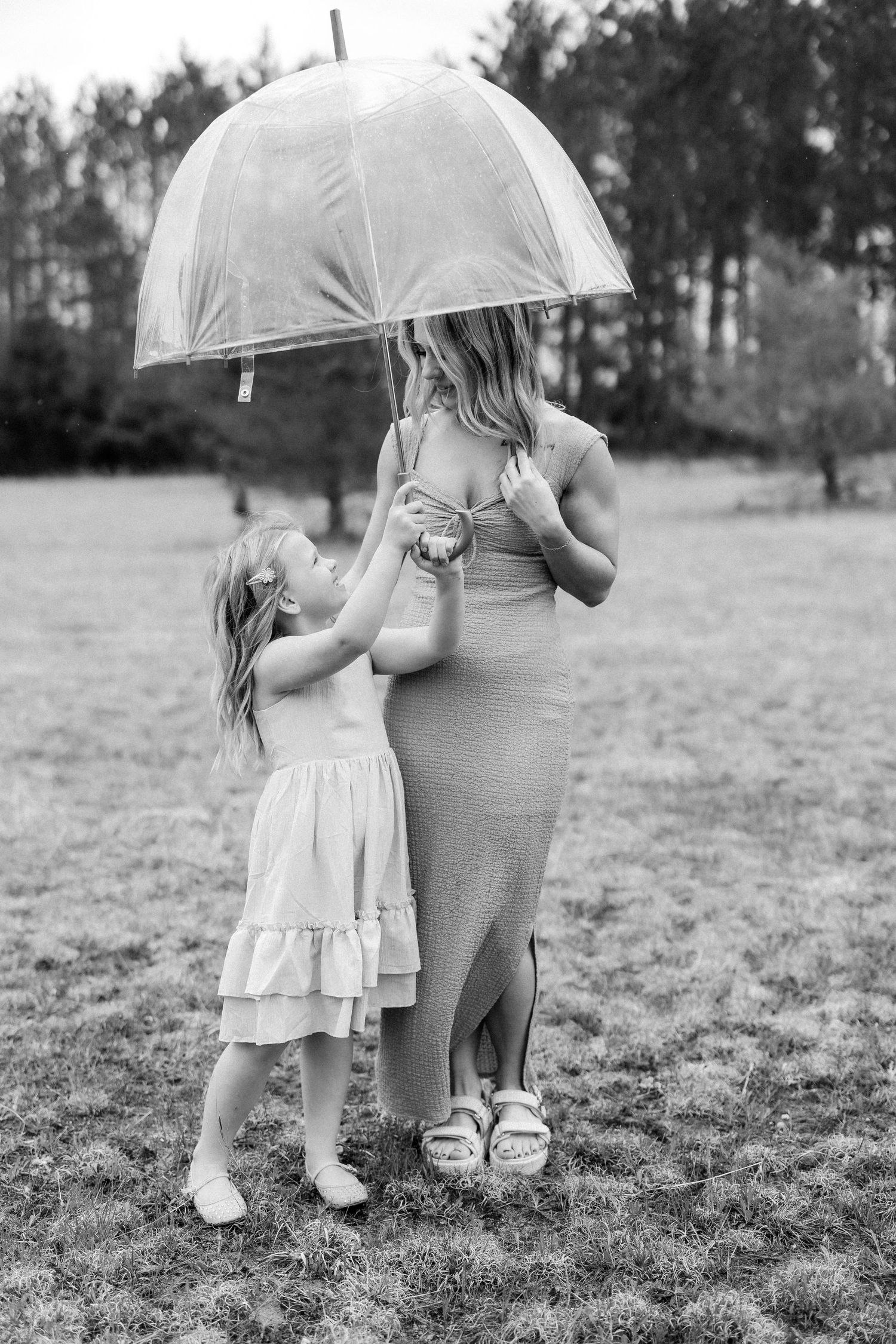 Black and white photo of a pregnant woman and young girl sharing a clear umbrella outdoors in a grassy field.