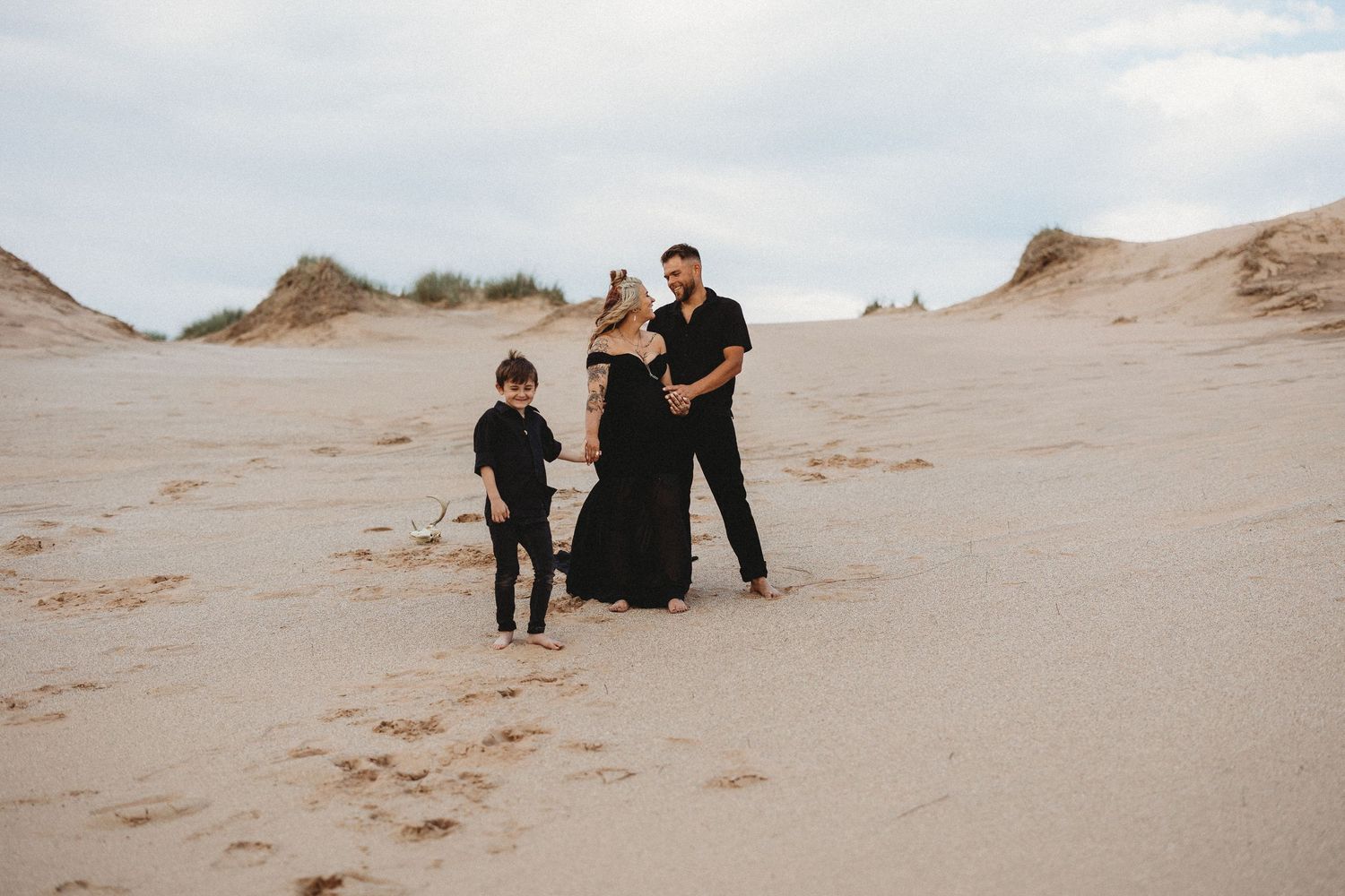 A family wearing black outfits stands together on sandy dunes under an overcast sky.