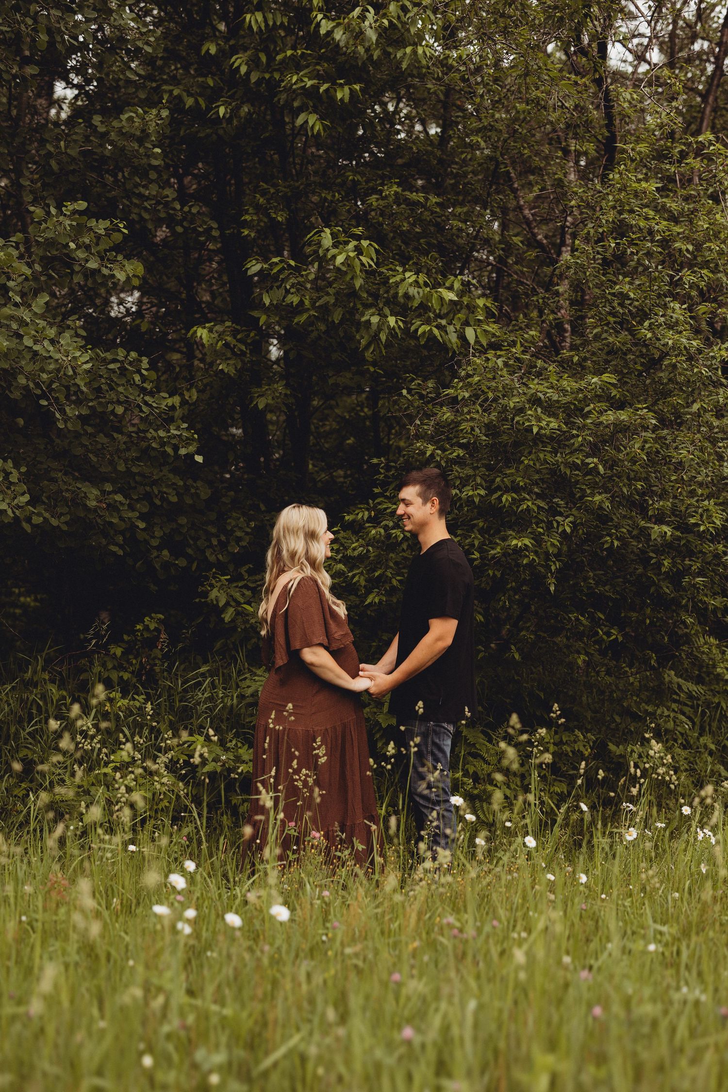 Couple holding hands in a wildflower meadow with lush green trees, woman in brown dress, man in black shirt.