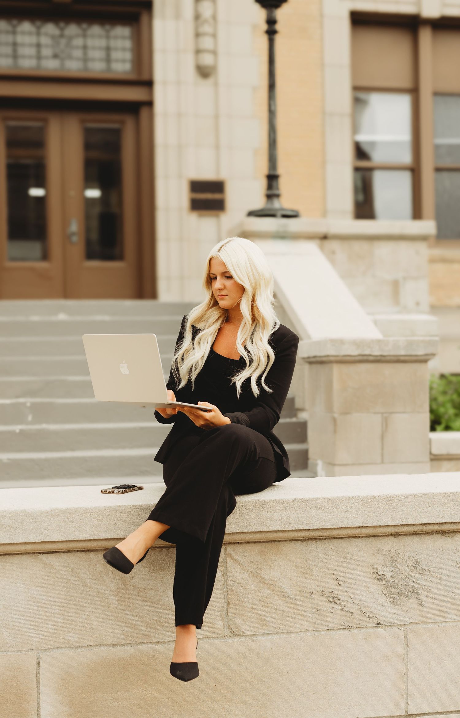 Blonde woman in black blazer and pants sits on stone steps with laptop outside classic building.