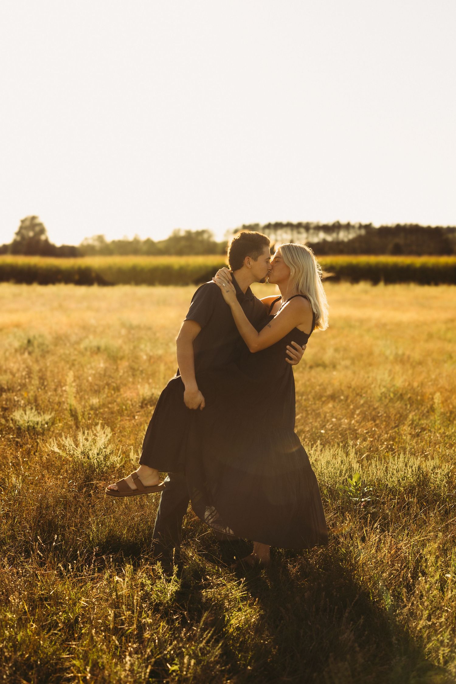 Couple kissing in a golden grass field at sunset, the woman in a long dark dress lifting one leg as the man holds her.