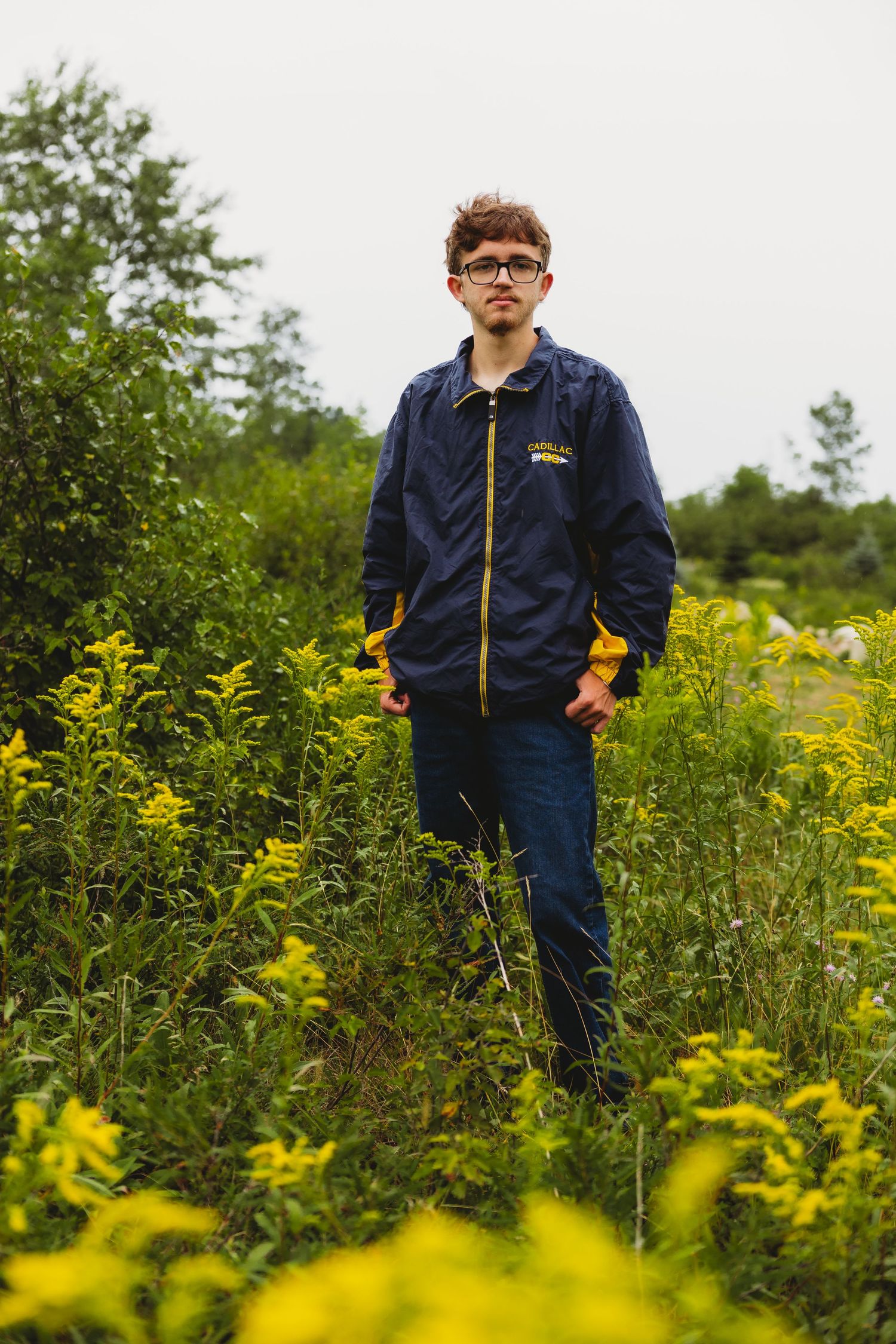 Young man with glasses and a navy jacket stands among blooming yellow goldenrod wildflowers in a green field under an overcast sky.