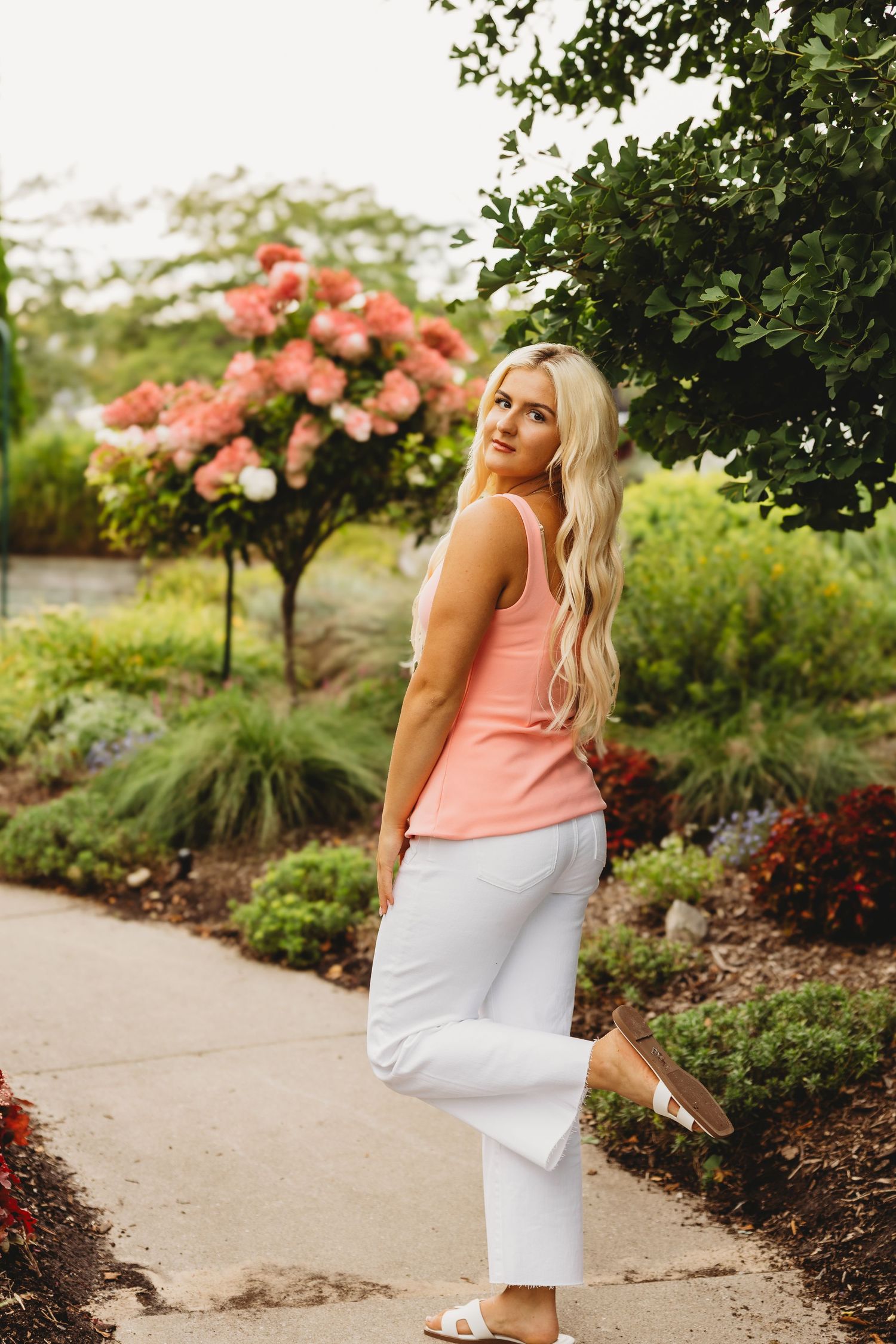 Woman with long blonde hair in a coral tank top and white pants stands on a garden path near blooming pink hydrangeas.
