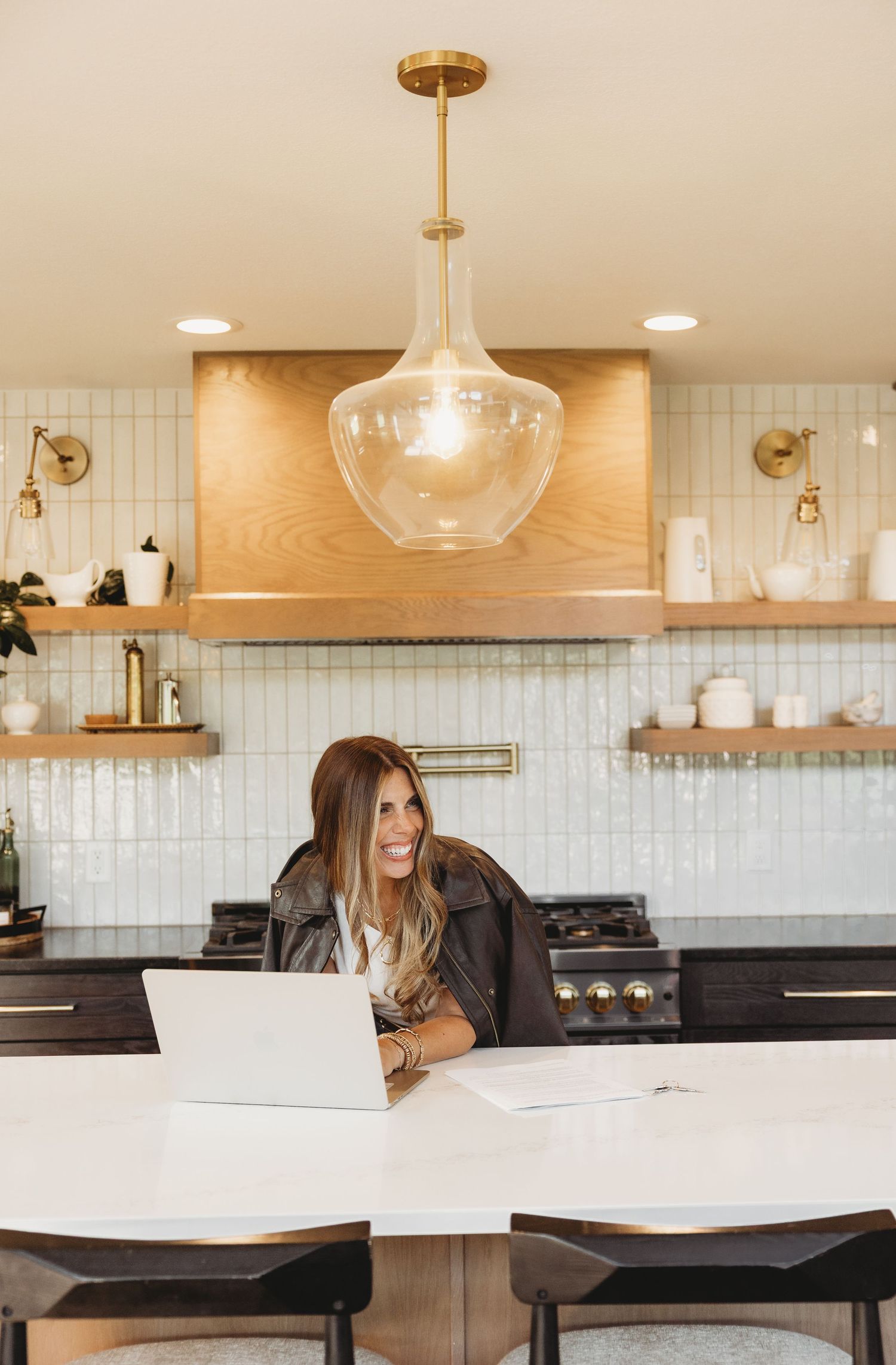 Smiling woman in a leather jacket works on a laptop at a white marble kitchen island beneath a glass pendant light and wood range hood.