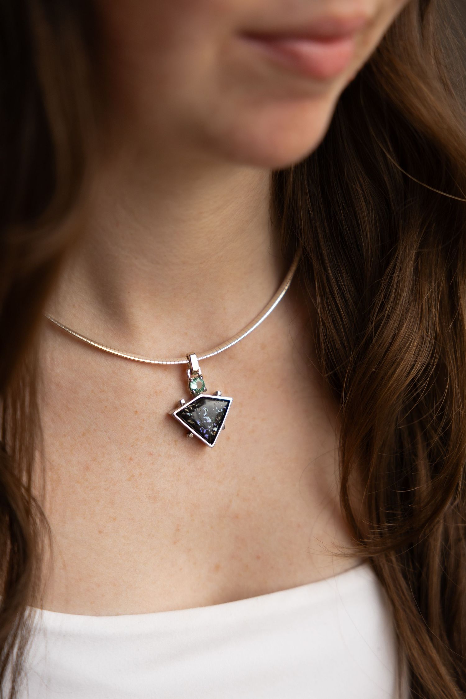 Woman wearing a sterling silver omega necklace with a triangular dark blue stone pendant topped by a small green gemstone.