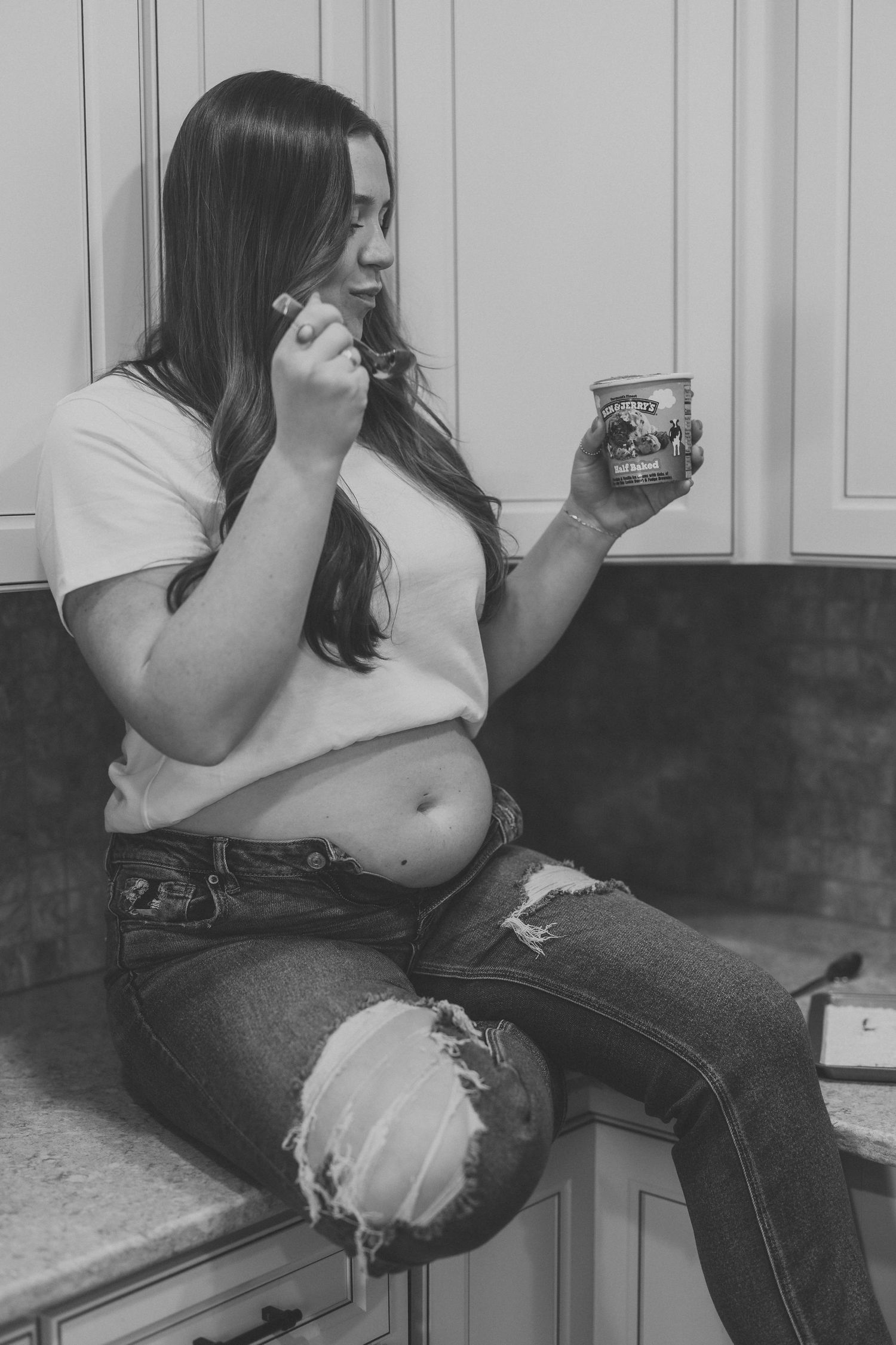Woman sitting on kitchen counter eating ice cream from a pint container in black and white photo.