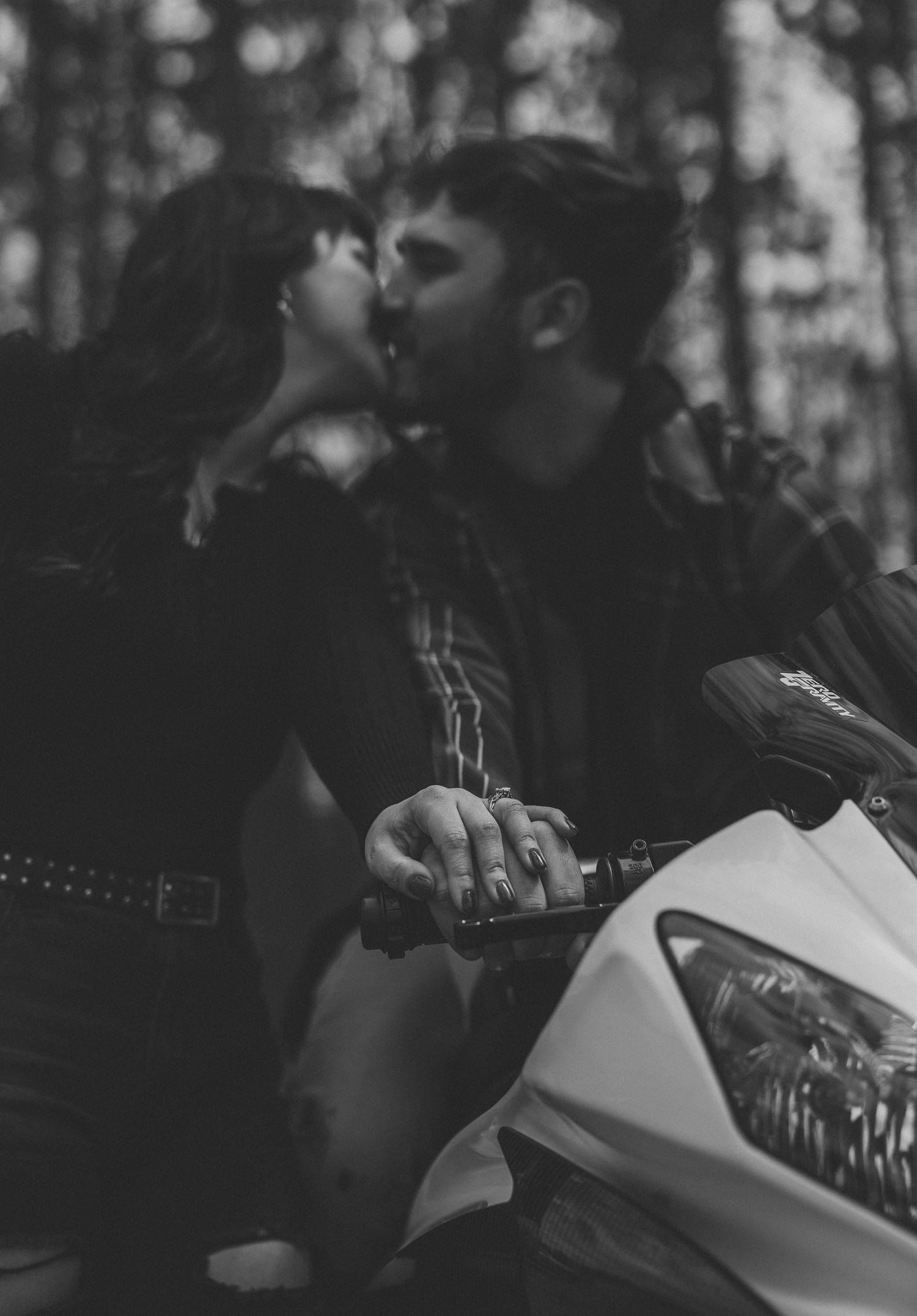 A couple shares a romantic kiss beside a white motorcycle in a forest, captured in black and white photography.