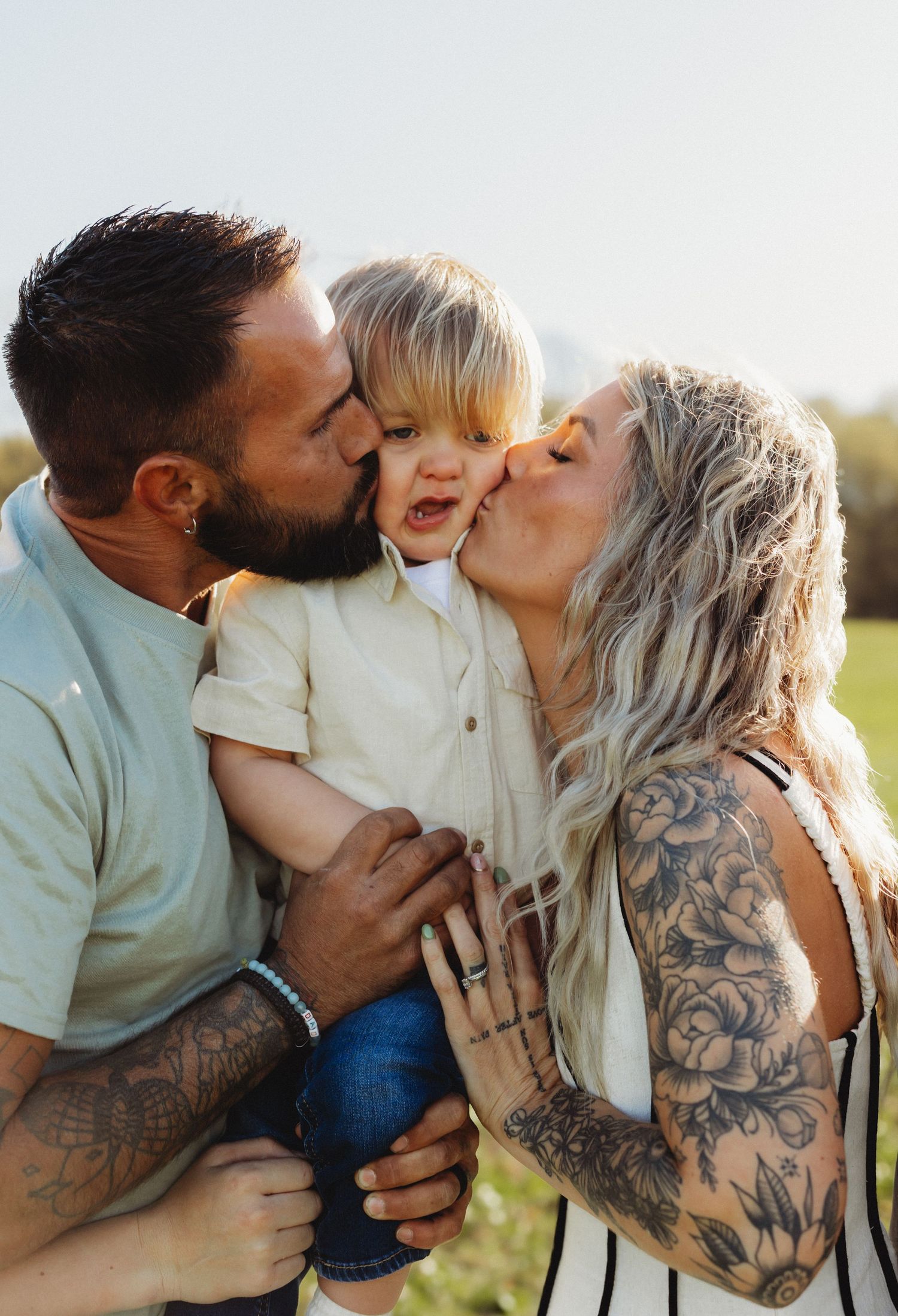 Tattooed parents kissing their smiling toddler on the cheeks in a sunny outdoor field.