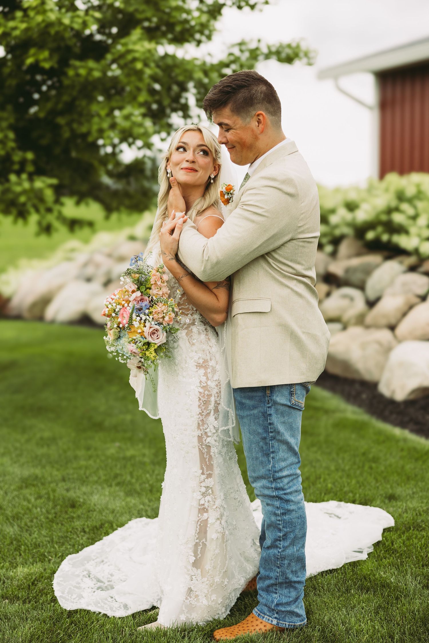 A romantic wedding portrait on a lush lawn with a rustic barn and landscaped garden in the background.