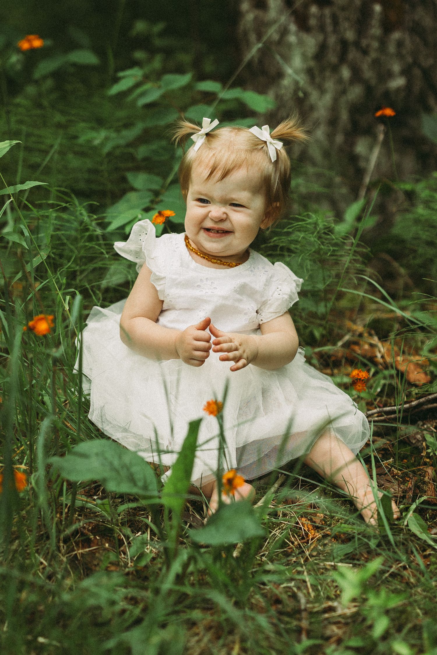 A baby in a white lace dress sits among orange wildflowers and green foliage in a garden setting.