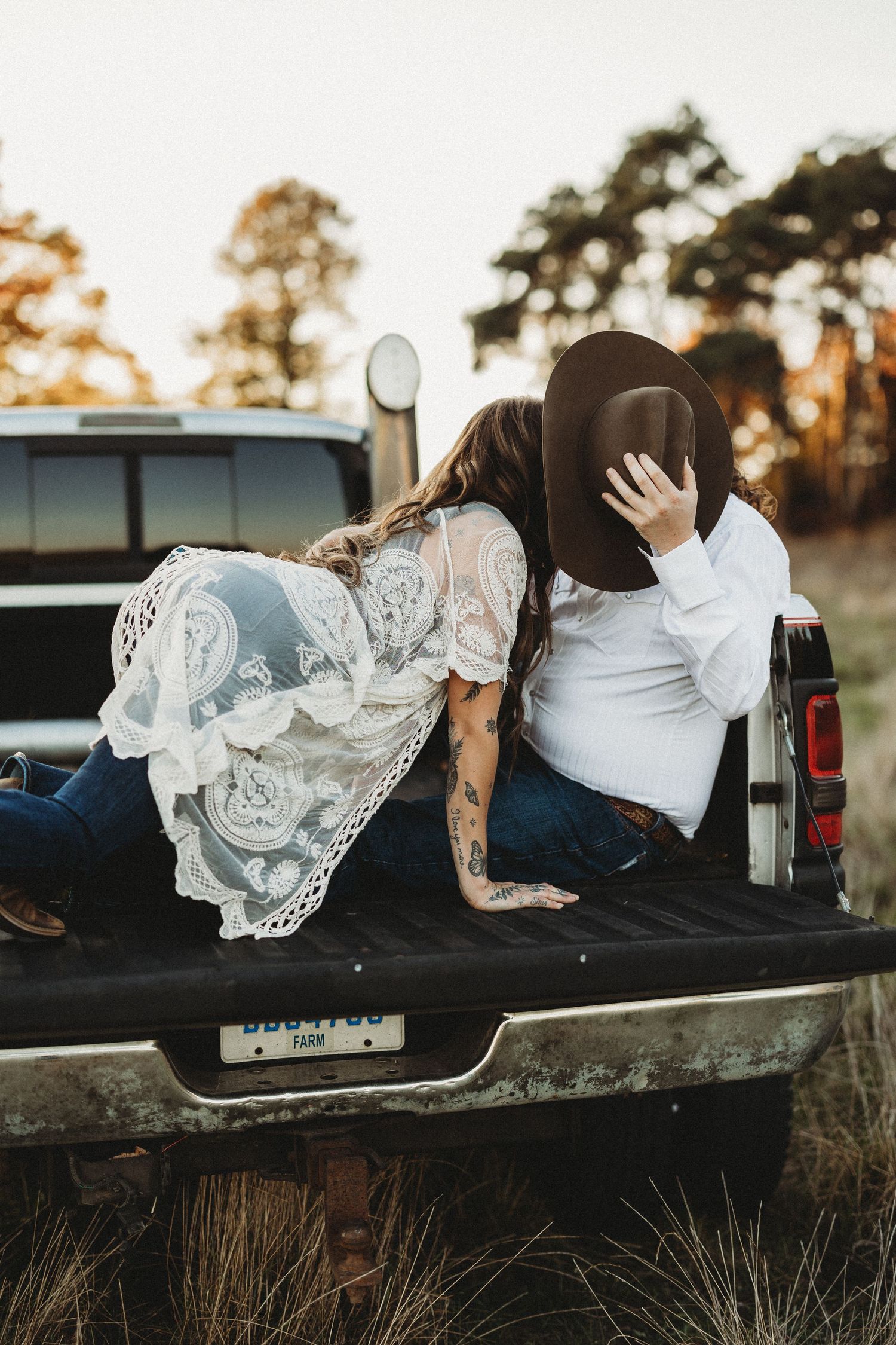 A couple in wedding attire share a romantic moment while lying in the back of a pickup truck at sunset.
