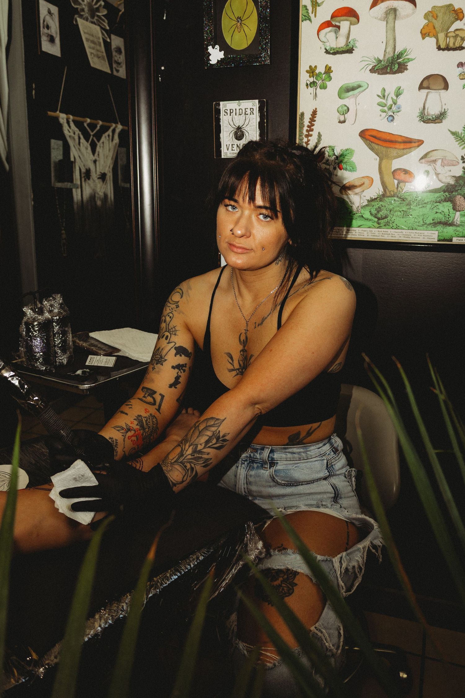 Tattooed woman in black crop top and denim shorts sitting in a tattoo studio with mushroom poster on wall.