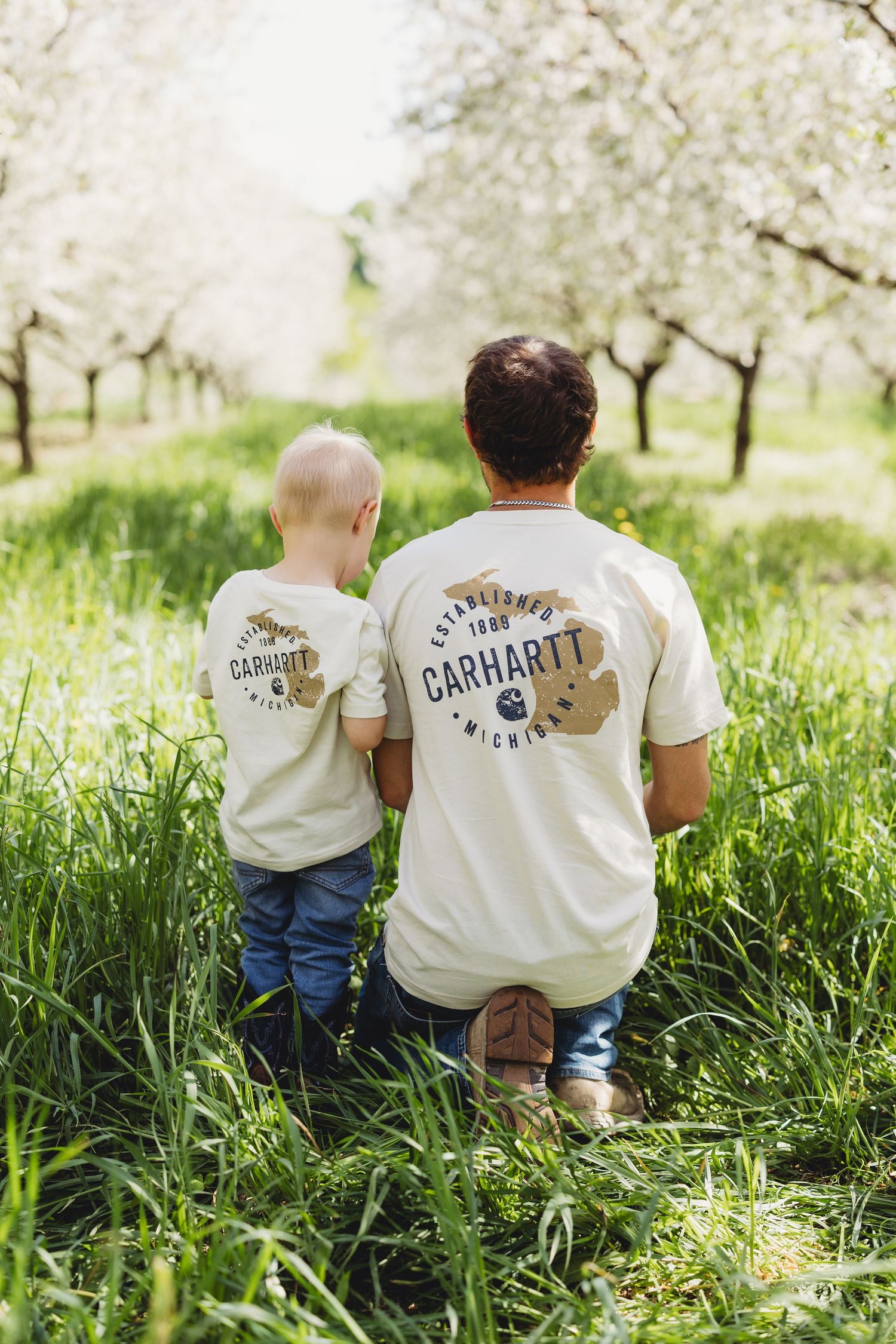 A father and young son wear matching Carhartt t-shirts while kneeling together in a blossoming spring orchard.