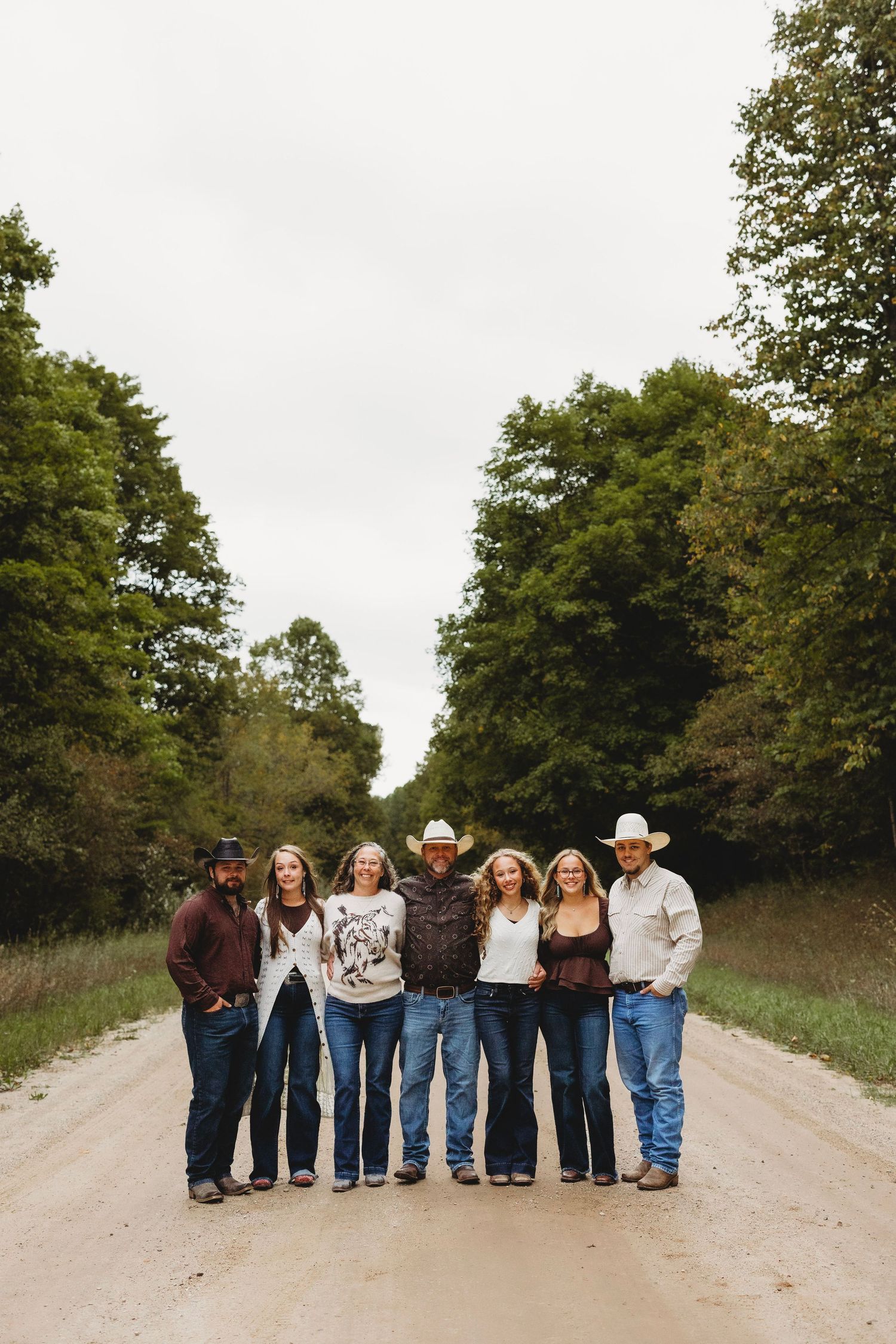Family of seven in jeans and cowboy hats standing arm in arm on a gravel country road lined with green trees under a cloudy sky.