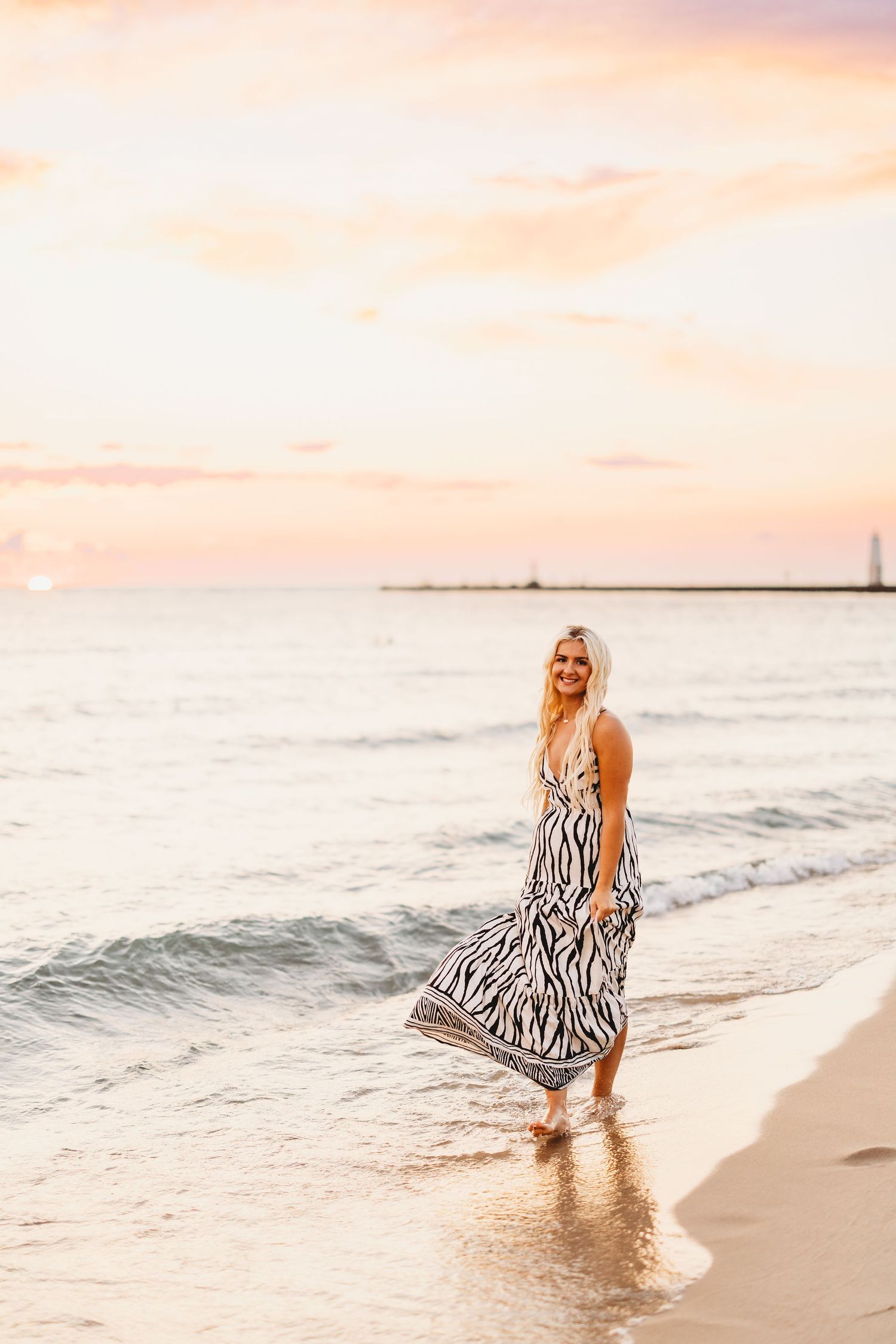 Woman in a black and white zebra print maxi dress walking barefoot along the shoreline at sunset with a pier in the distance.