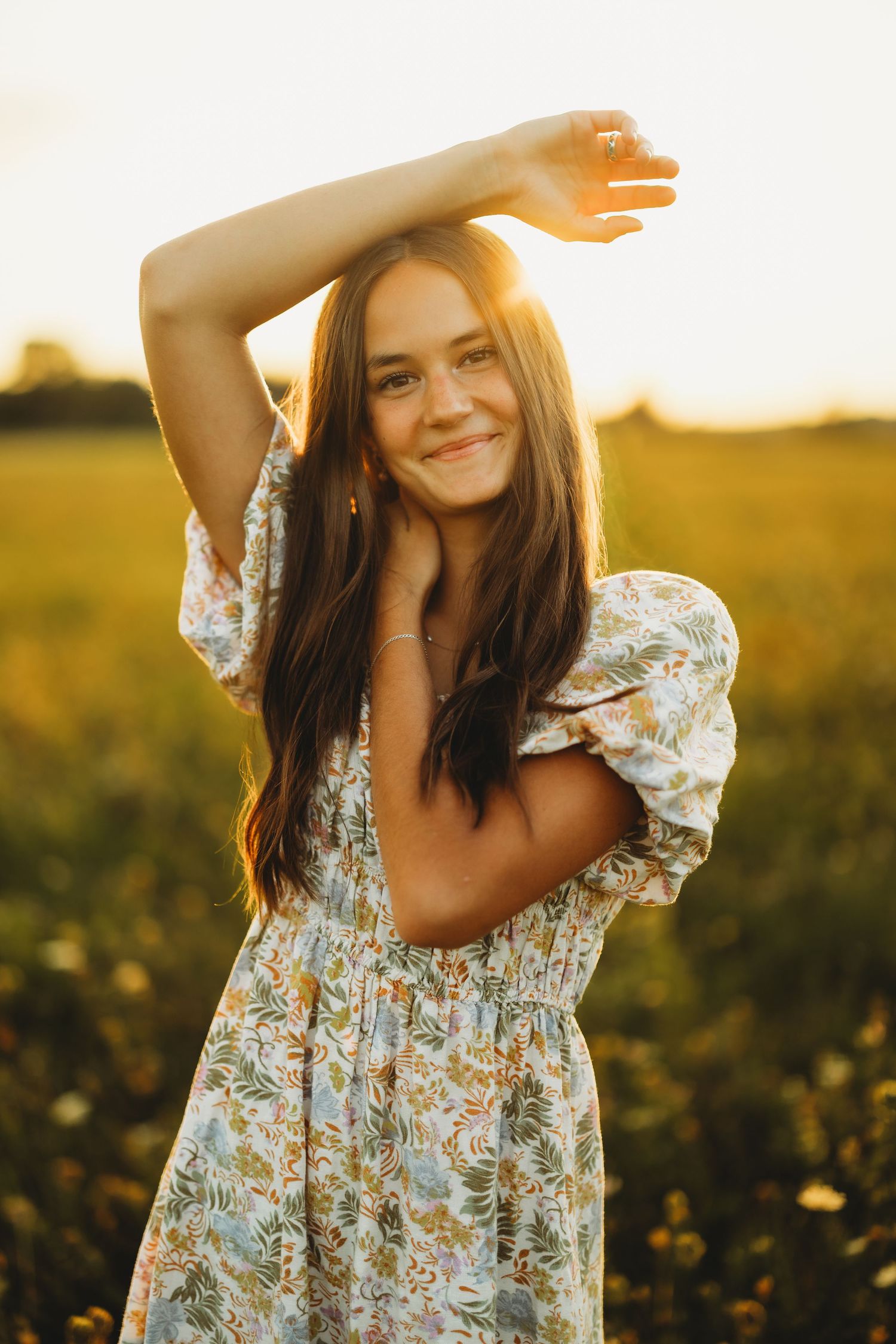Smiling young woman with long brown hair in a floral dress raises one arm overhead in a golden sunlit field at sunset.