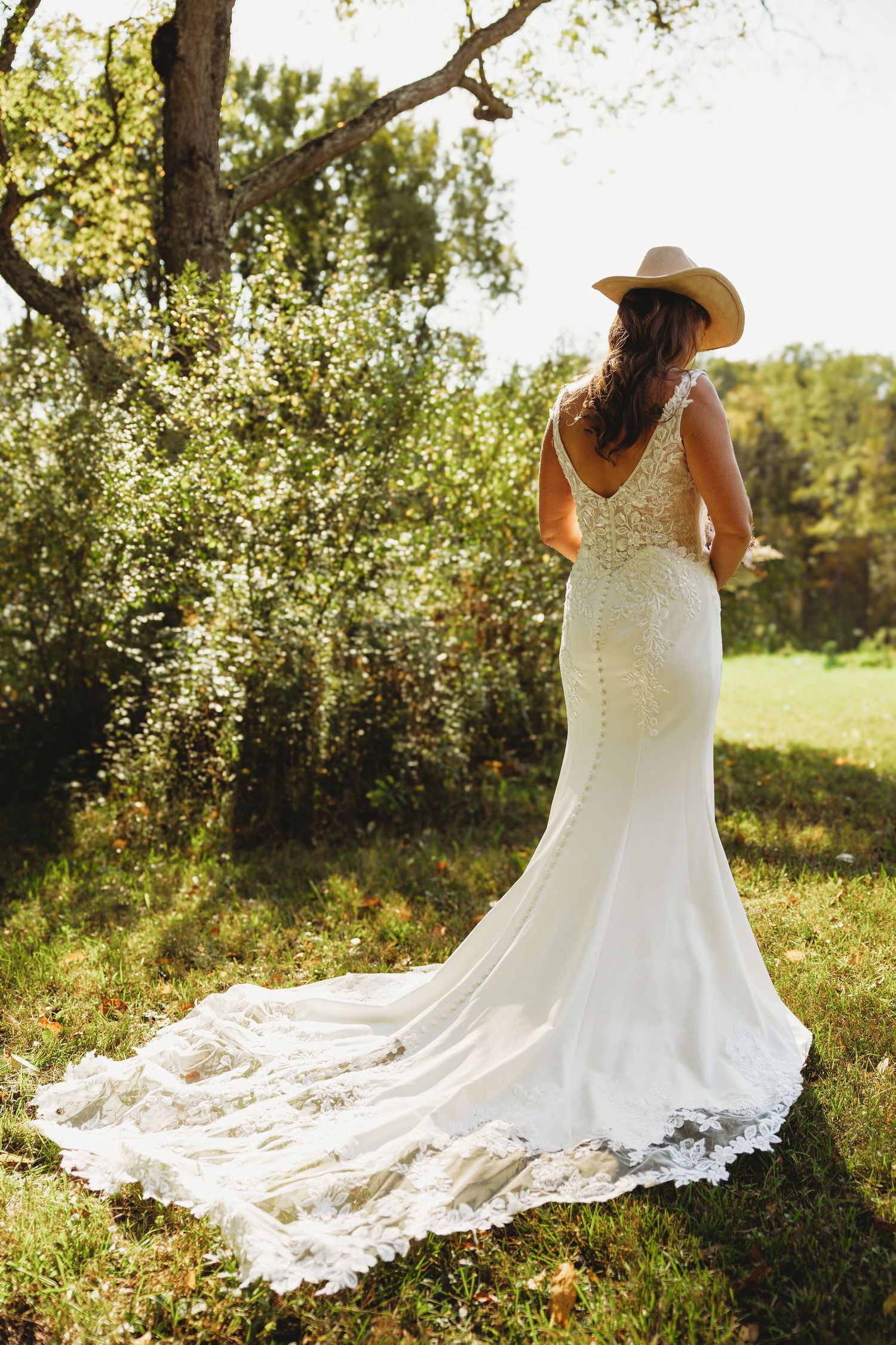 Bride in a lace mermaid wedding gown with a long train and a cowboy hat stands facing a sunlit grassy field and trees.