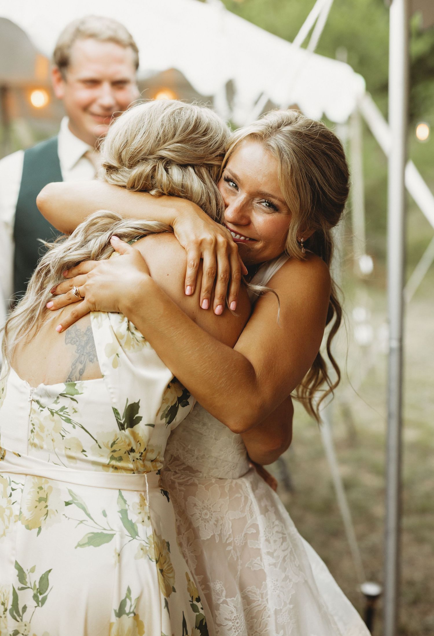 Two women in floral and lace dresses share a warm hug at an outdoor tented wedding while a smiling man looks on.