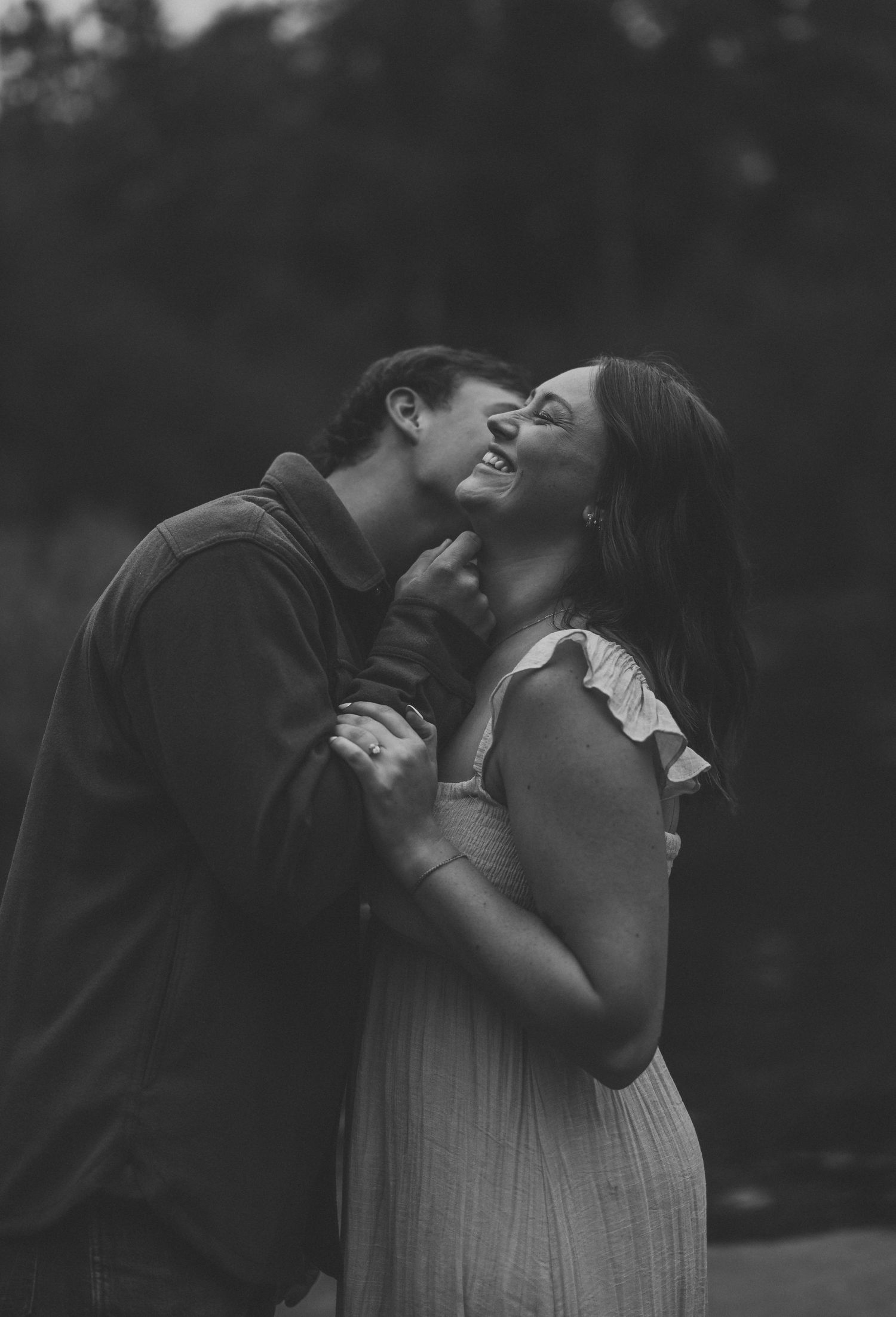 Black and white photo of a smiling couple embracing outdoors as a man kisses the cheek of a laughing woman in a ruffled dress.