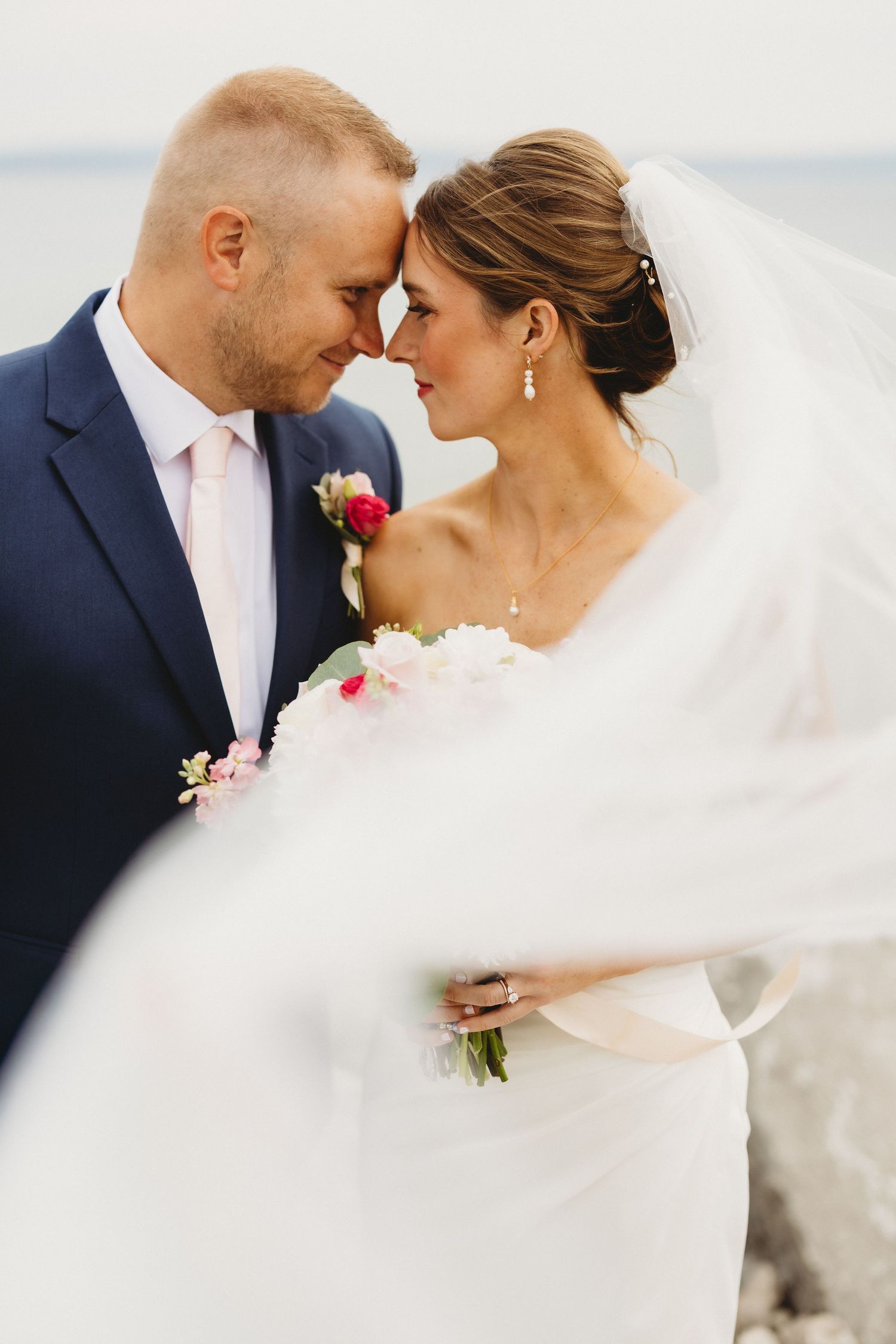 A bride and groom touch foreheads romantically outdoors, she holds a pink bouquet, her veil flowing in the wind.