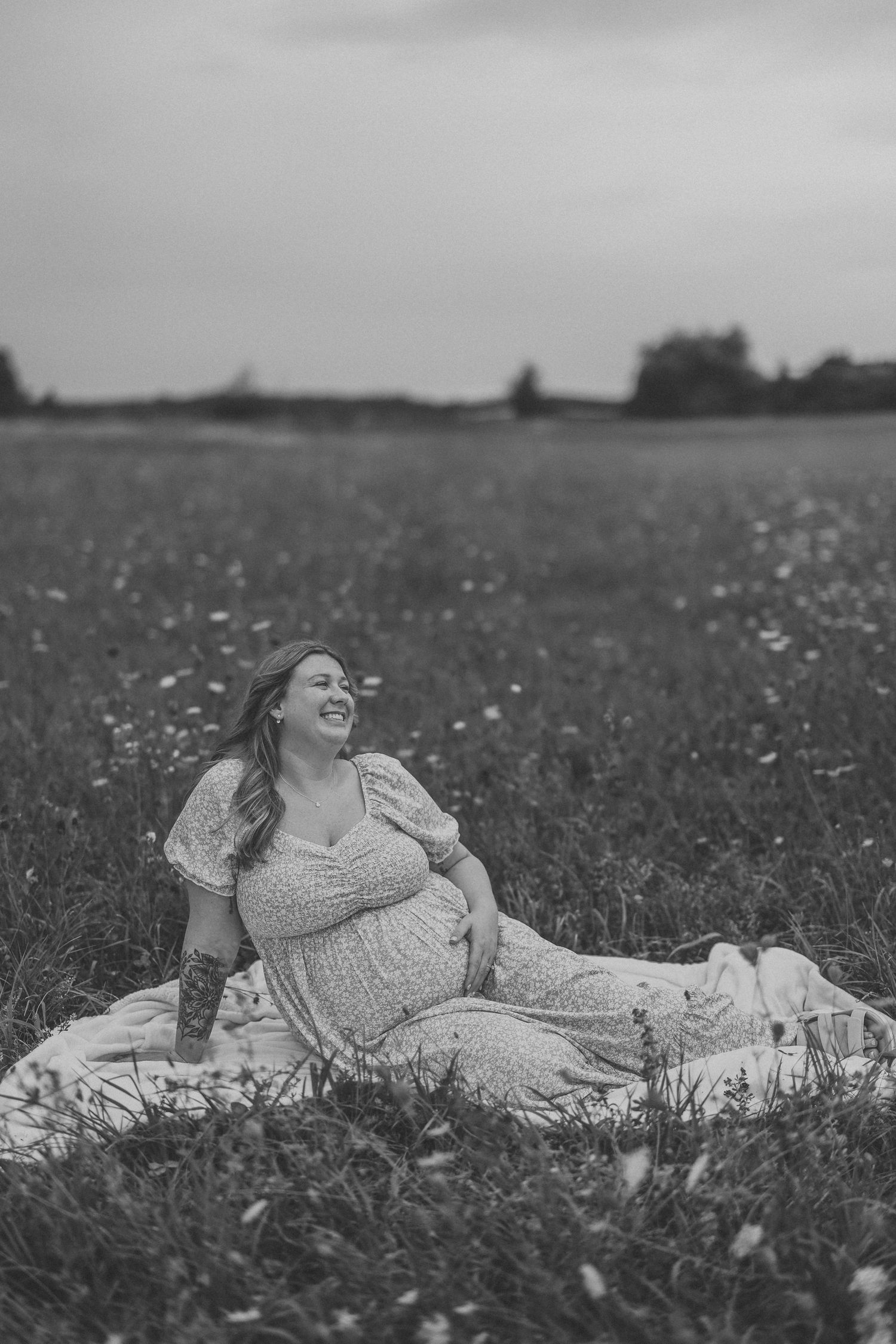 Black and white photo of a smiling pregnant woman in a floral dress sitting in a meadow field.