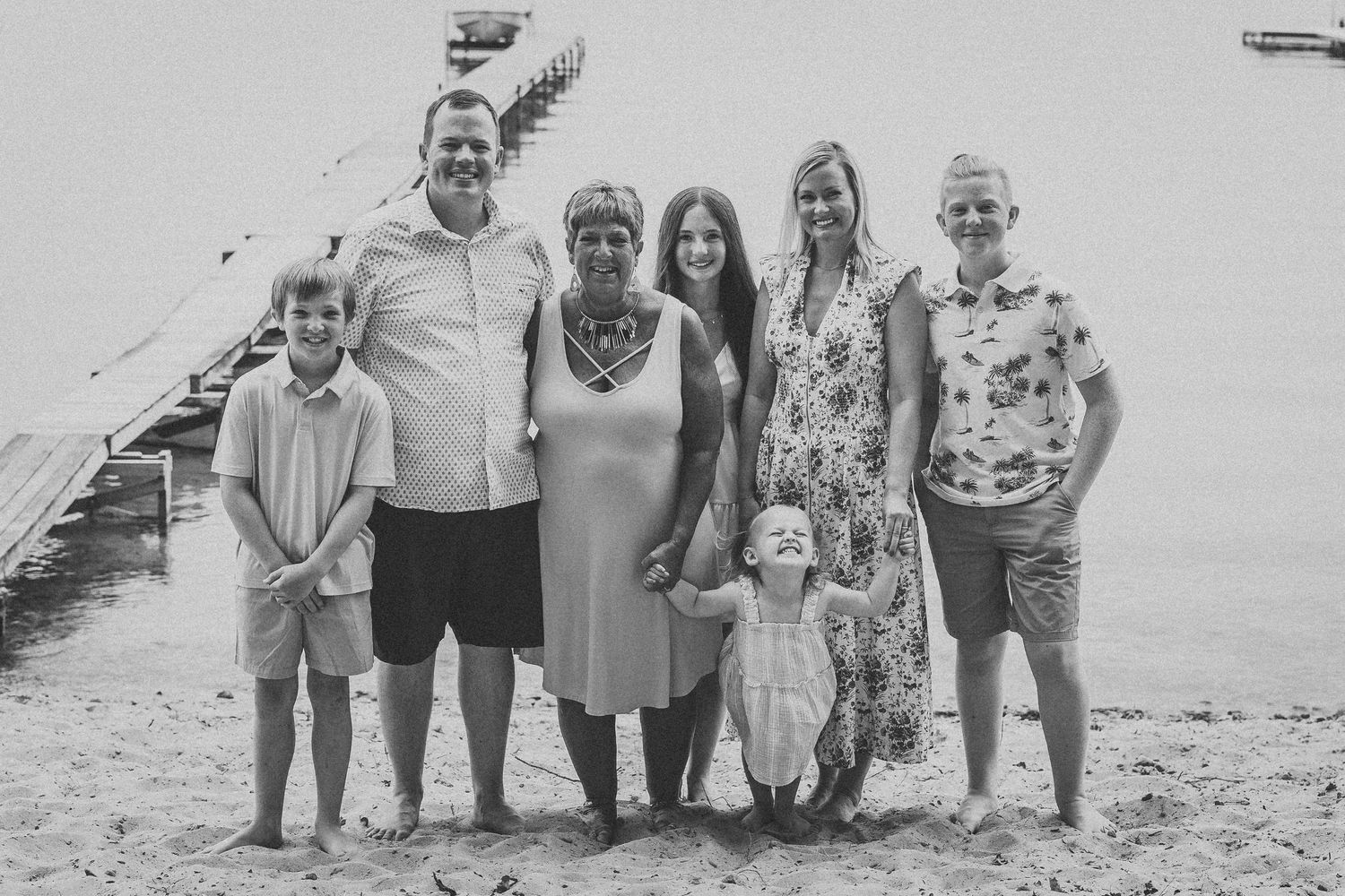 Black and white family portrait of seven people posing together on a sandy beach with a pier in the background.