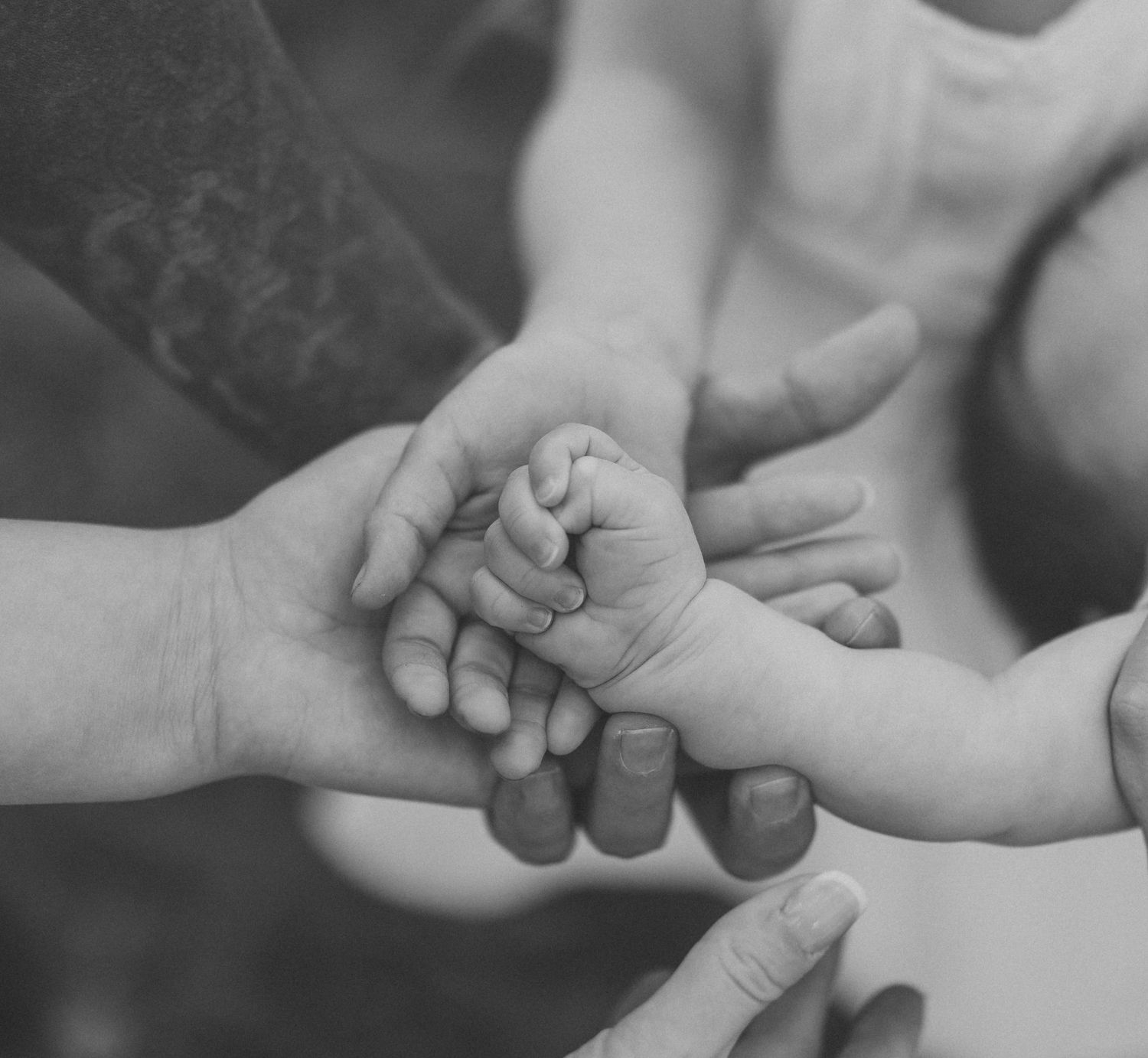 Black and white photo of multiple hands coming together, including a tiny newborn baby hand held by adult hands.