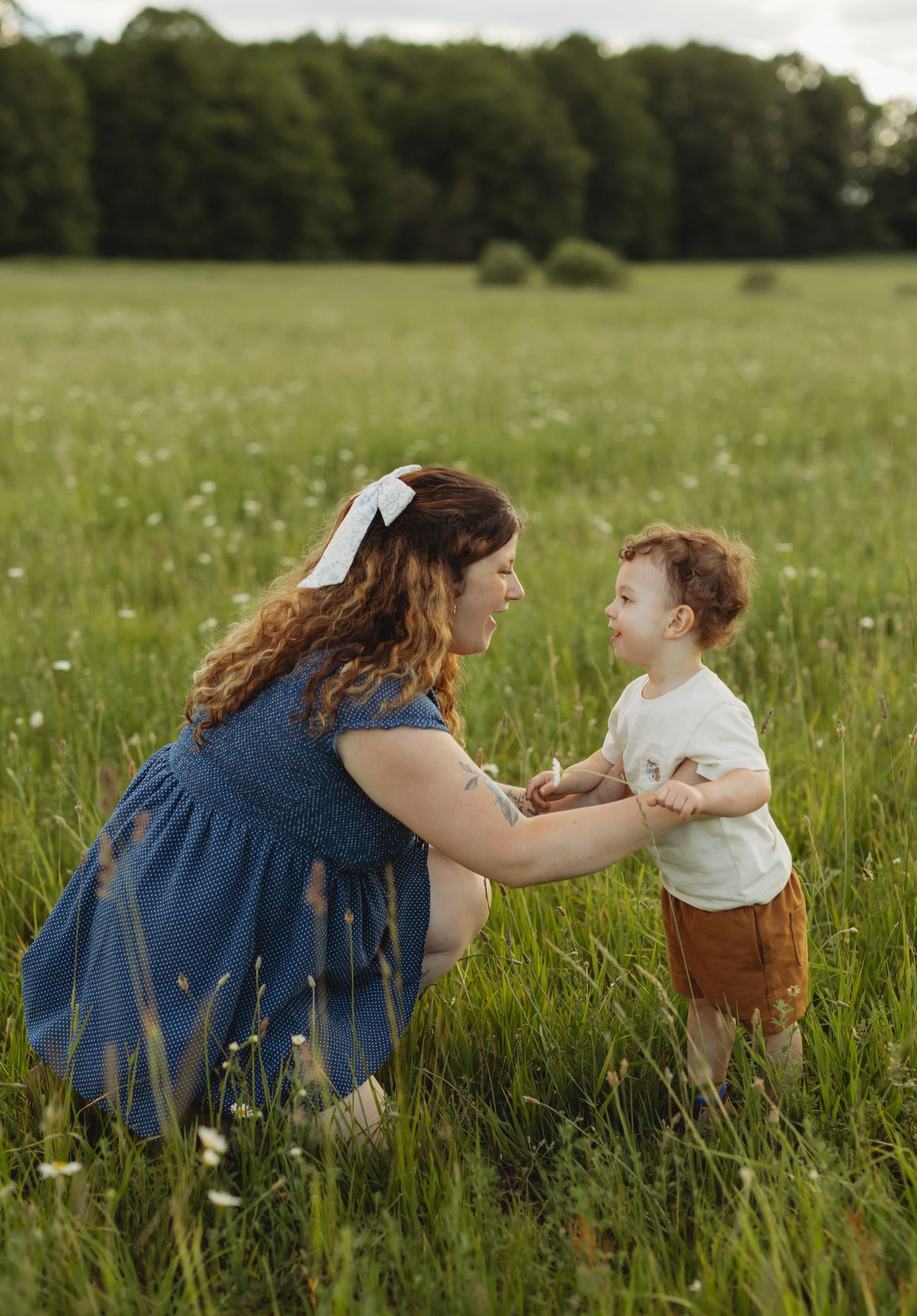 A mother in a blue dress kneels in a green field, holding hands with her toddler son on a sunny day.