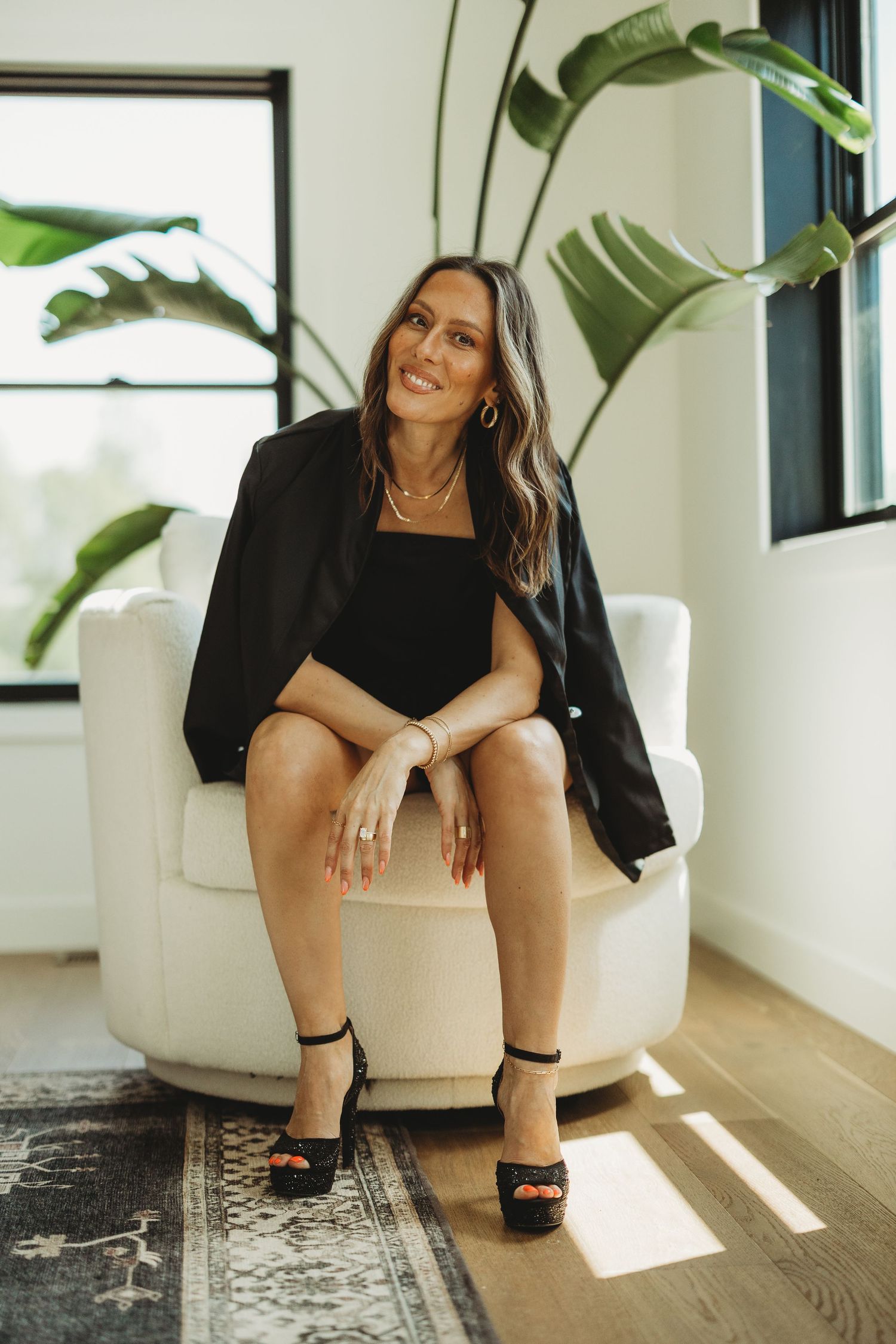 Woman in black blazer and heels sitting in white chair with tropical plants in bright modern interior.