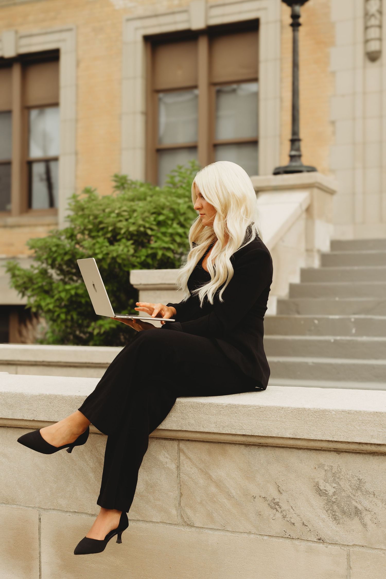 Blonde woman in black suit sitting on stone ledge outdoors, working on laptop near building steps.