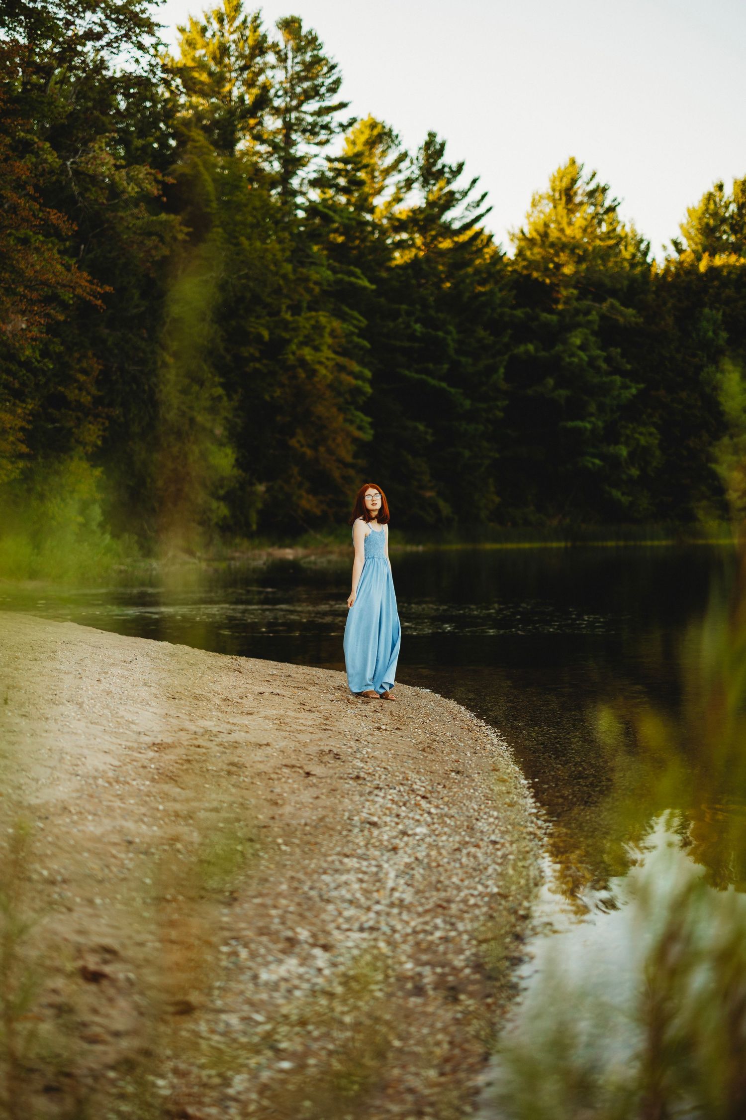 Woman in a flowing blue dress stands on a pebbly lakeshore with tall evergreen trees reflected in the calm water at sunset.