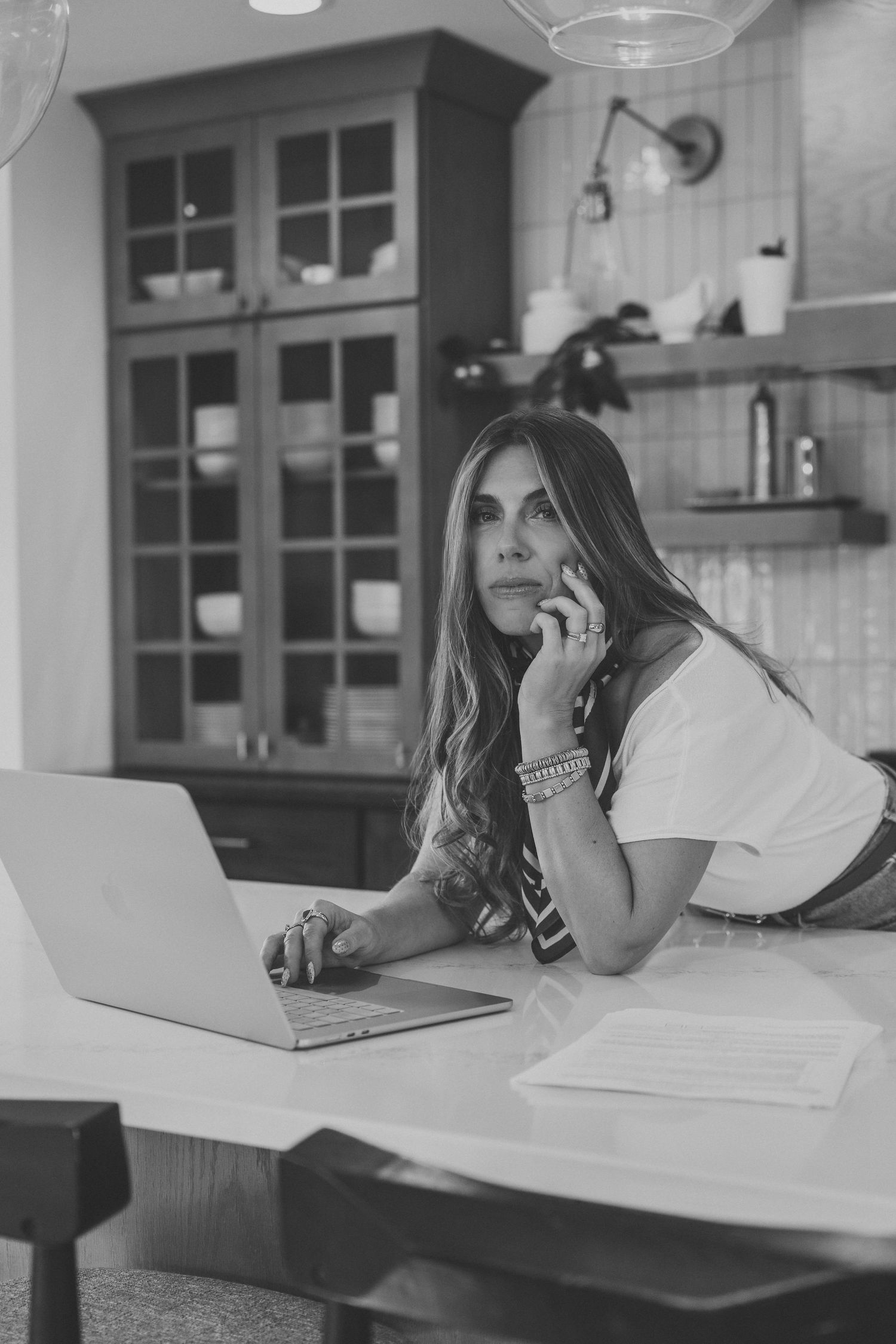 Woman with long hair talking on a phone while leaning on a kitchen island beside an open laptop, in a black and white photo.
