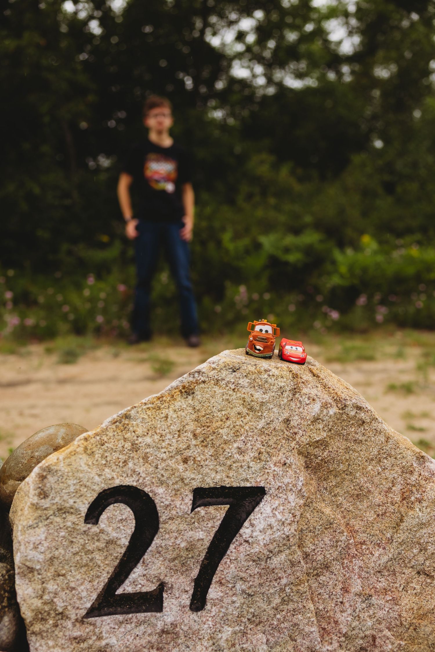 Two toy cars from the Cars movie sit atop a granite boulder engraved with the number 27 while a blurred boy stands in the background.