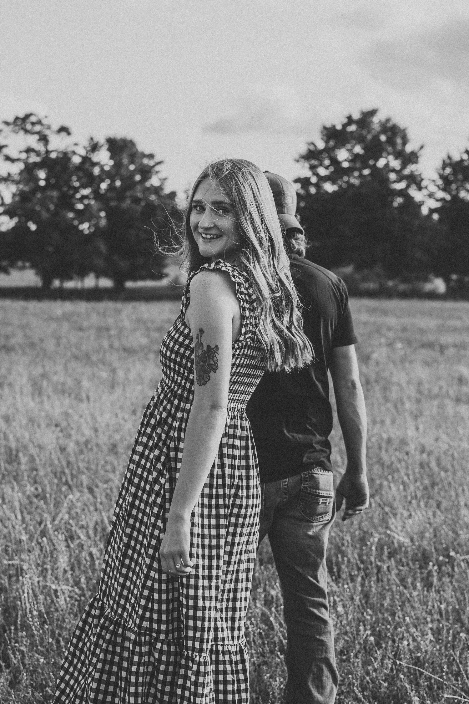 A couple in a grassy field shares a candid moment together, with her wearing a gingham dress and him in dark clothing.