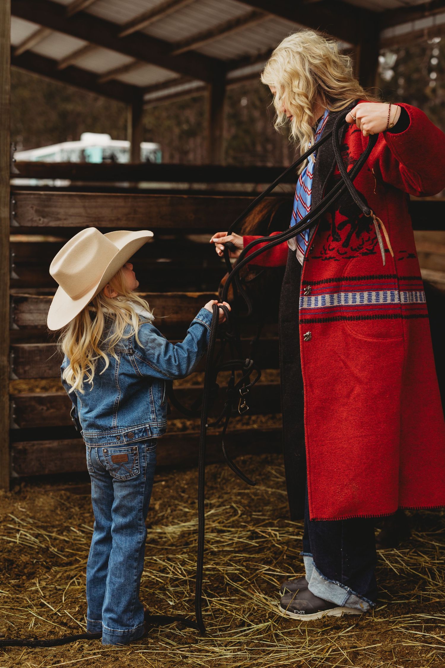 Children in cowboy boots and denim explore a rustic barn with hay-covered floors while wearing western attire.
