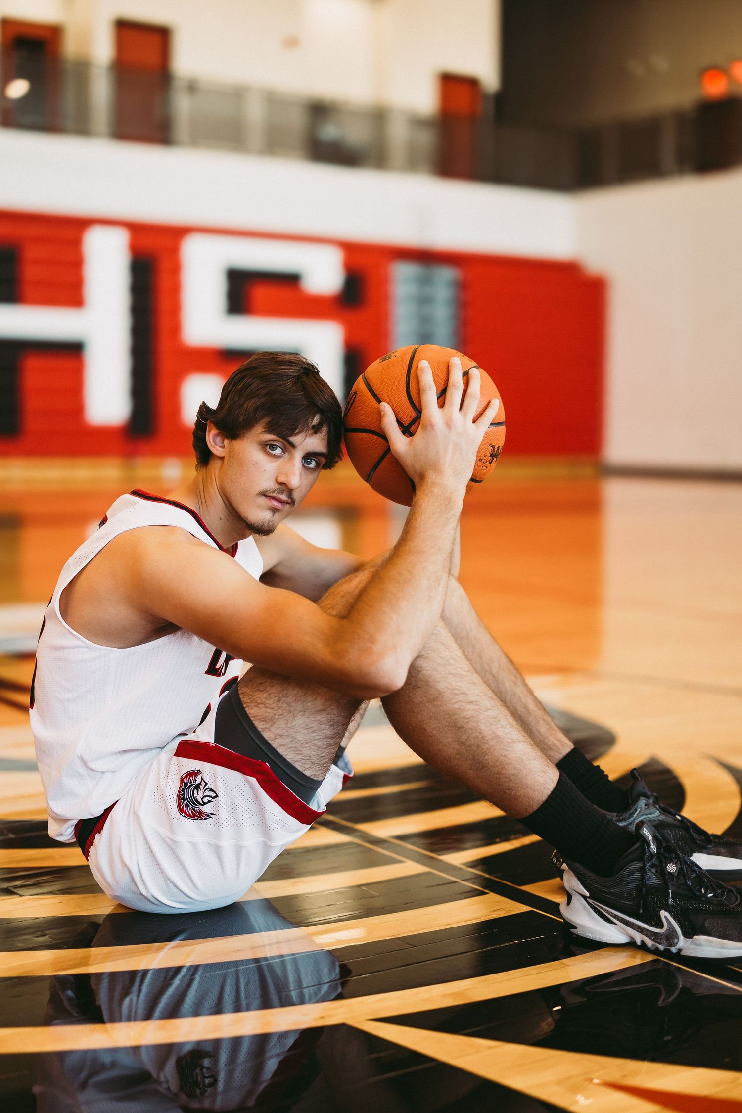 Basketball player in white uniform sits on court holding ball against red and black backdrop.