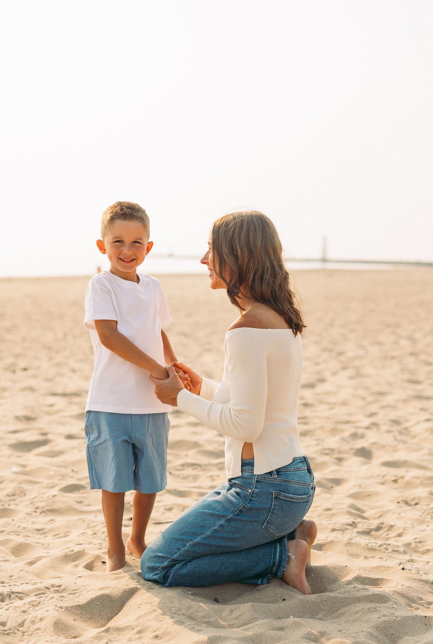 A woman kneels on sandy beach holding hands with a smiling young boy in white shirt and blue shorts.
