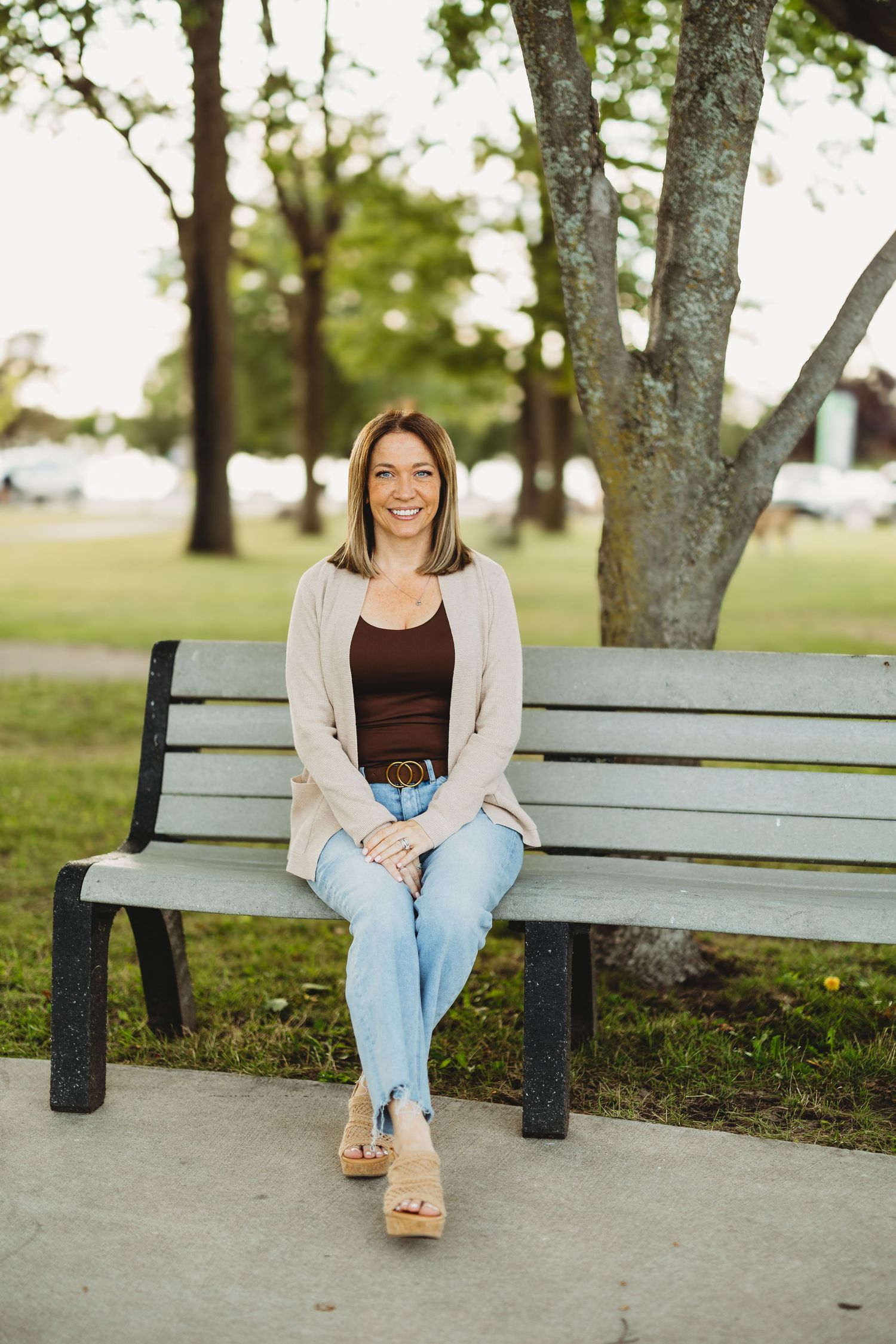 Smiling woman in jeans and cardigan sitting on a park bench surrounded by trees on a sunny day.