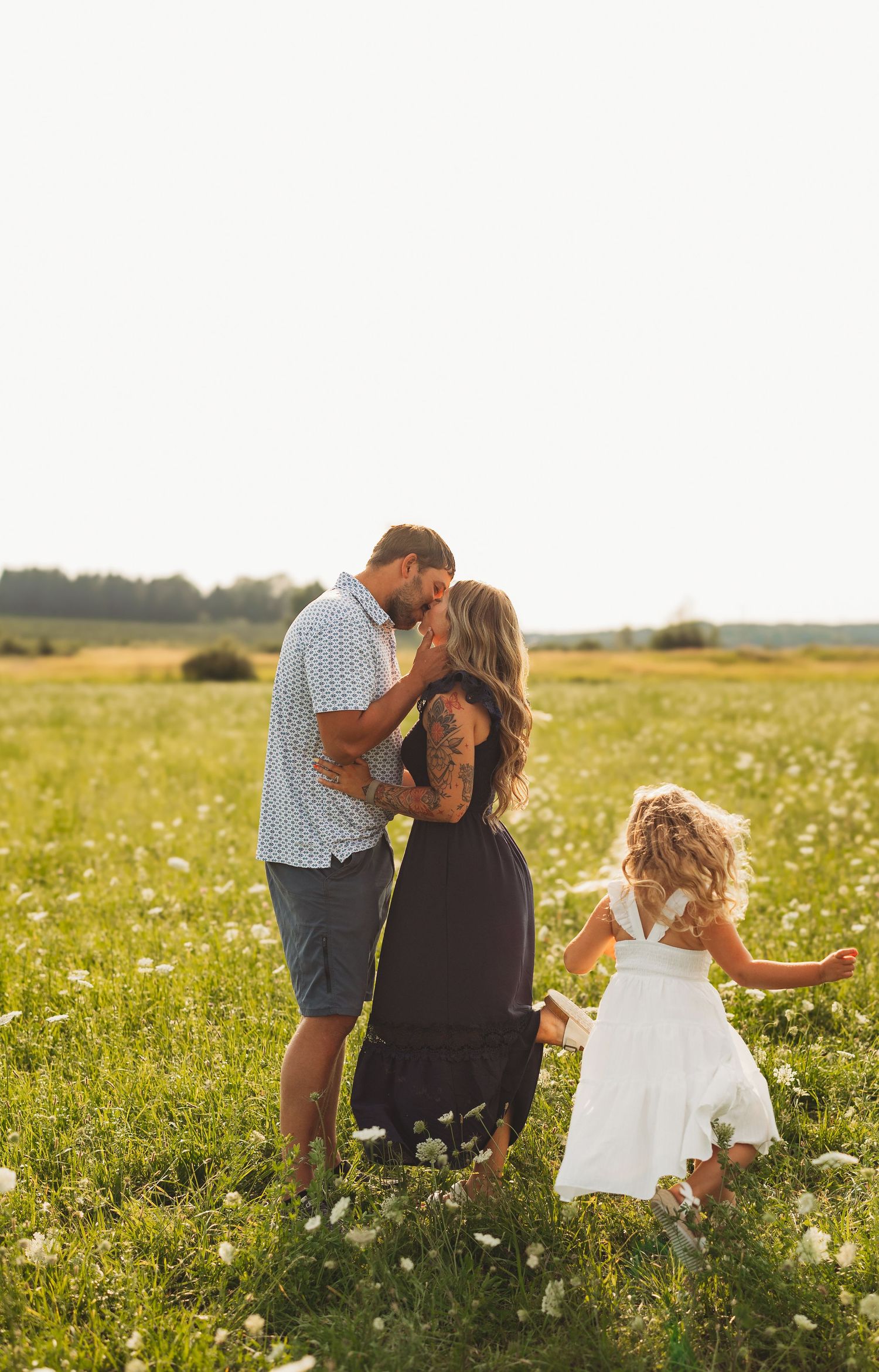 A couple shares a romantic kiss in a sunny wildflower field while their young daughter in a white dress plays nearby.