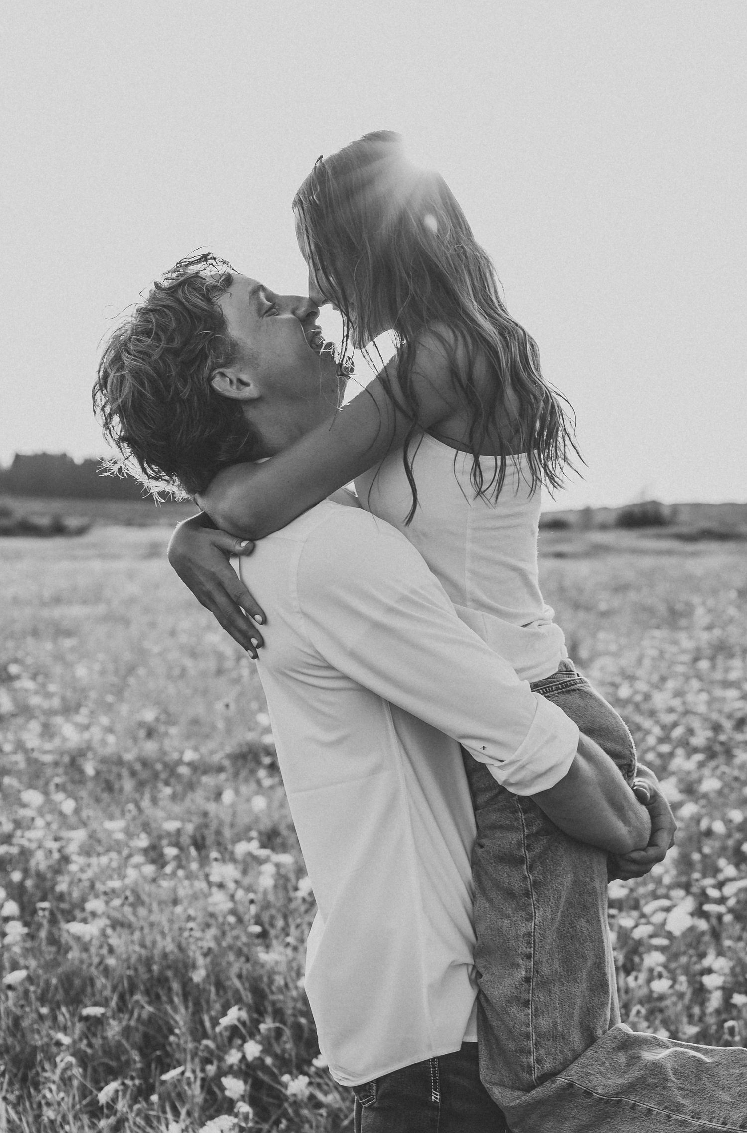 Black and white photo of a man lifting and kissing a woman in a sunlit flower field.