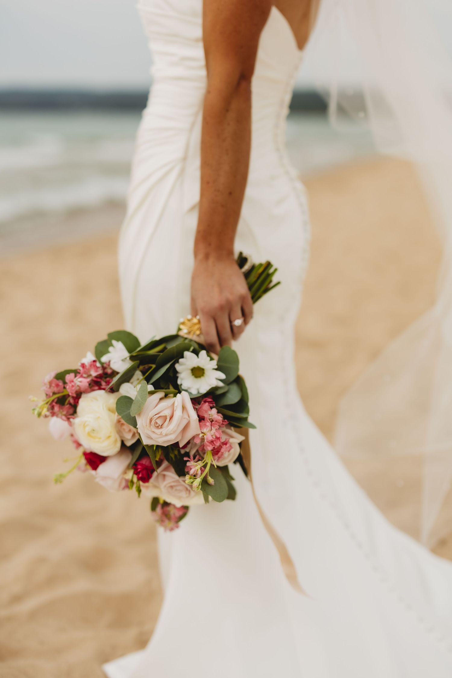 Bride in white gown holding pink and white floral bouquet on sandy beach with ocean backdrop.