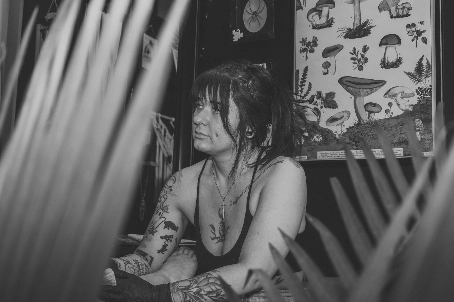 Black and white photo of a tattooed woman with dark hair sitting thoughtfully near palm leaves with mushroom art in background.