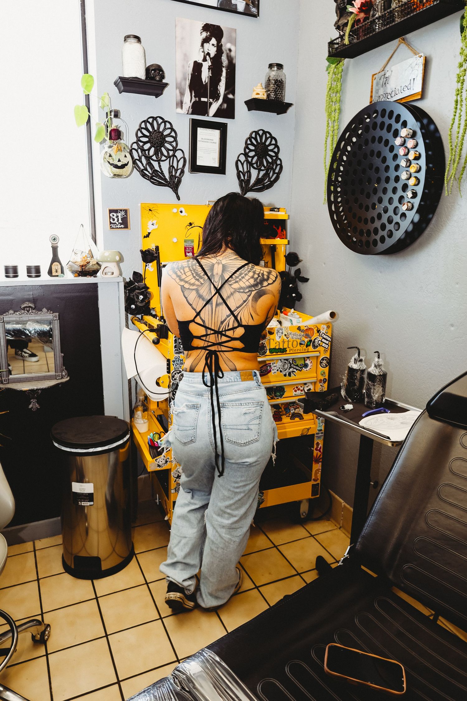 Tattooed woman with back tattoos standing at a yellow tool chest in a tattoo studio.