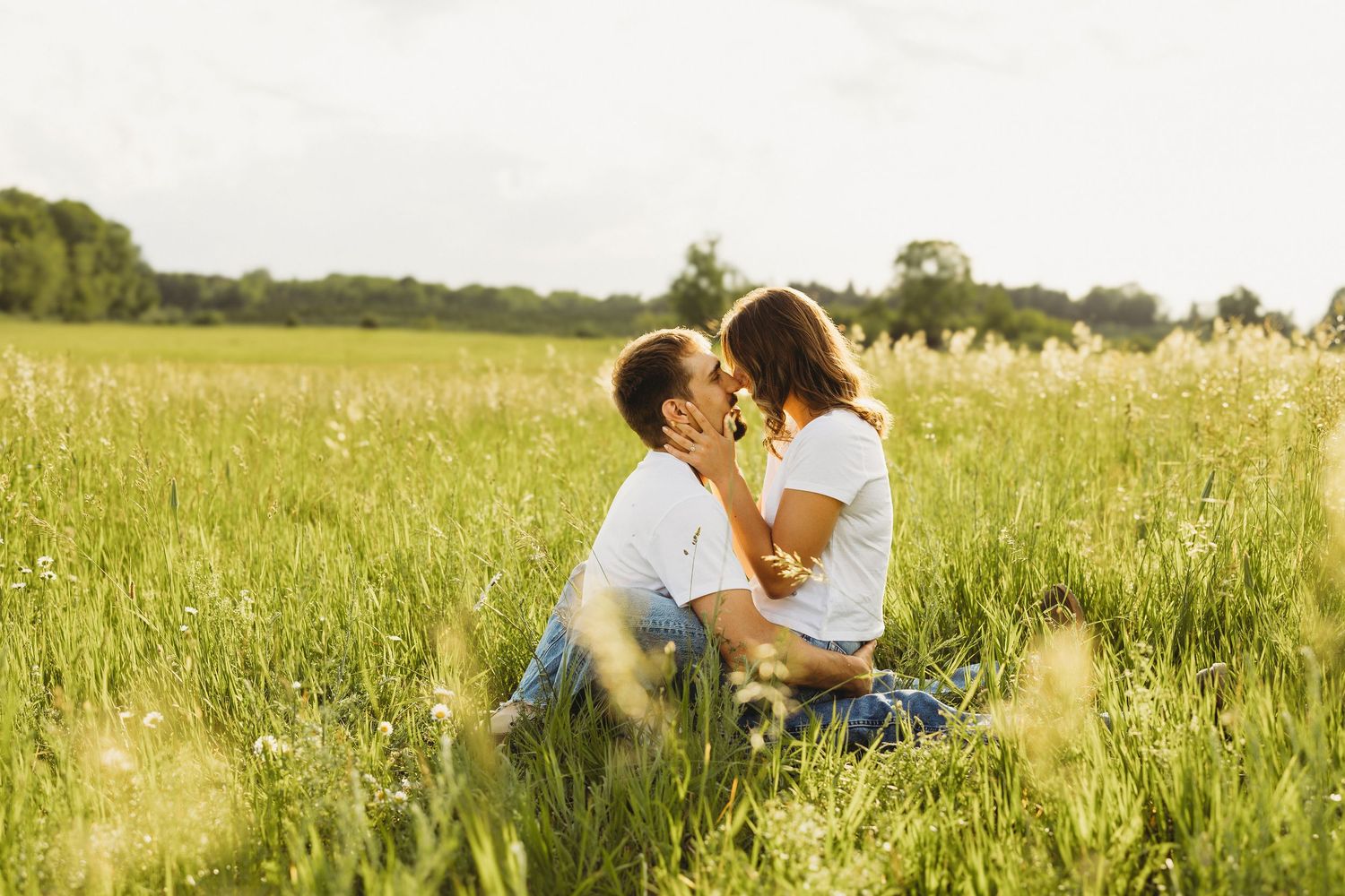 Romantic couple sitting in lush green meadow, leaning in for a kiss on a sunny summer day.