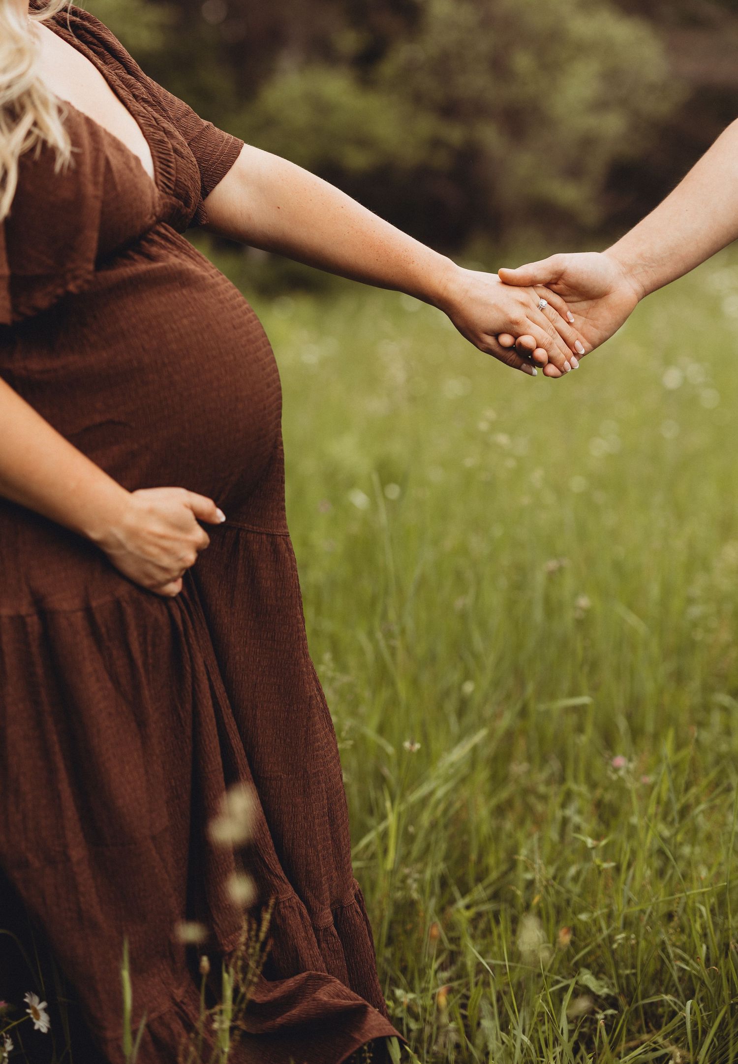 Pregnant woman in brown dress holding hands with partner in a lush green meadow during maternity photoshoot.