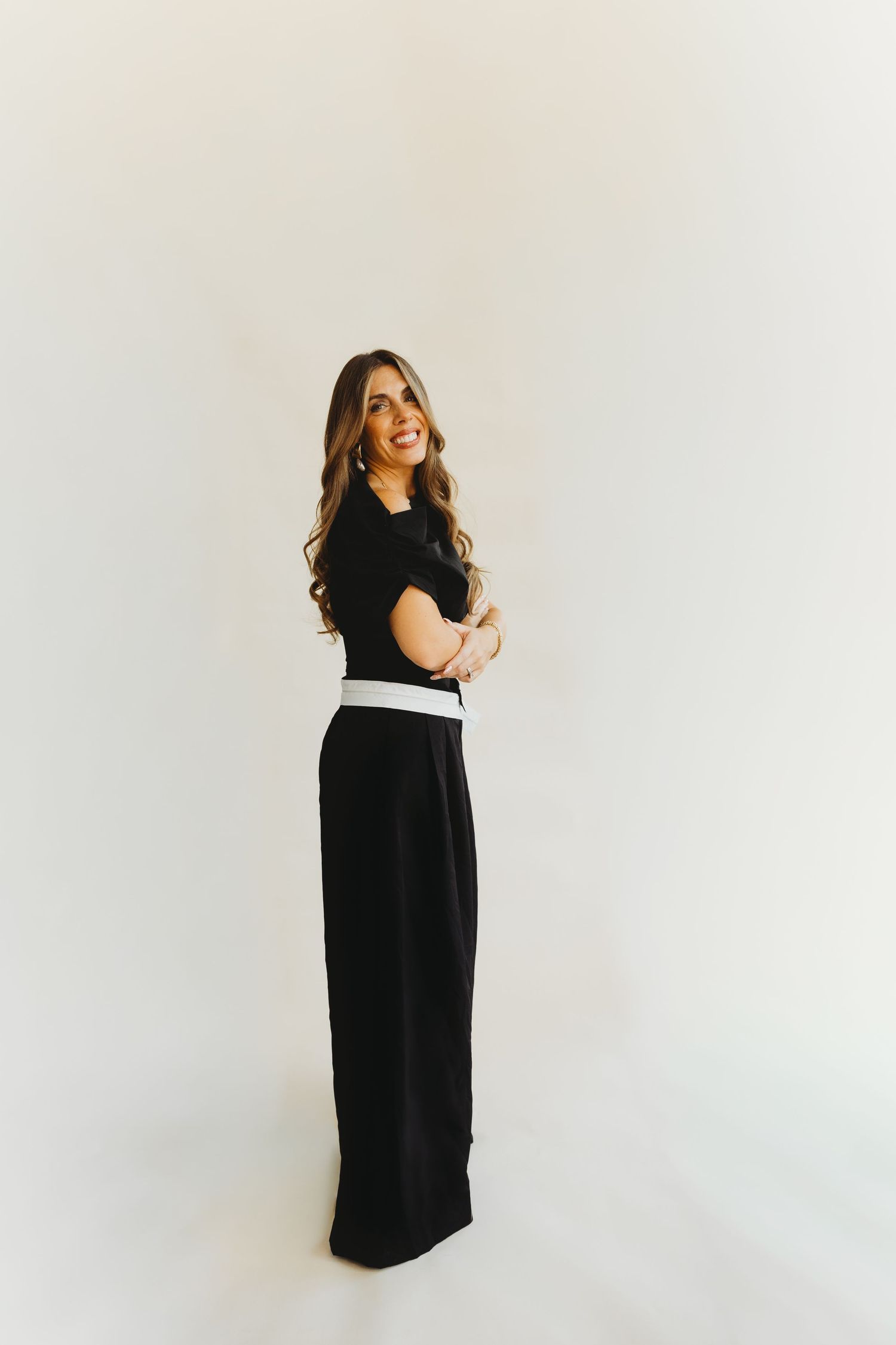 Smiling woman with long wavy hair poses sideways in a black short-sleeve top and wide-leg pants with a white belt against a plain backdrop.