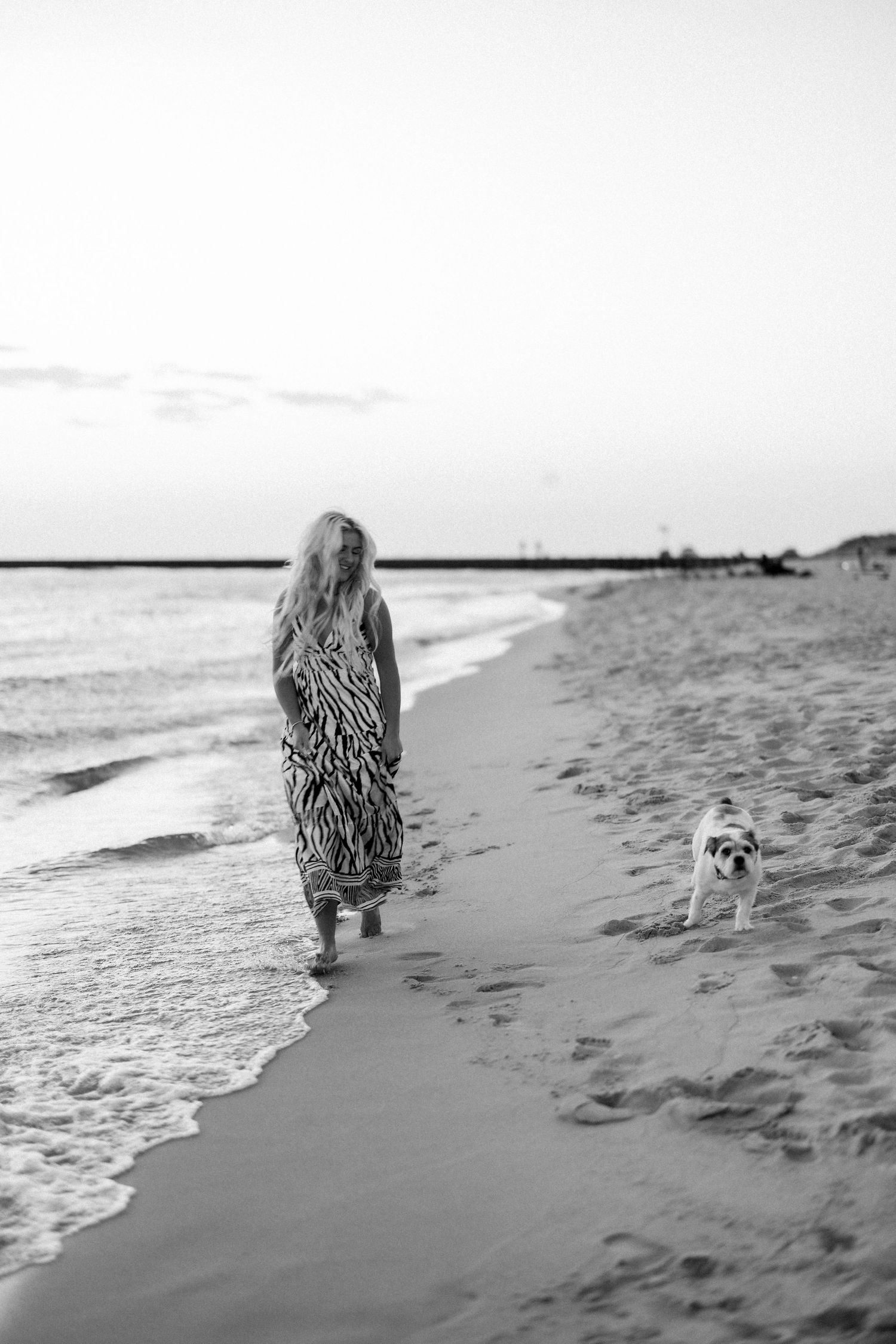 Black and white photo of a woman in a patterned dress walking along the beach shoreline with a small dog.