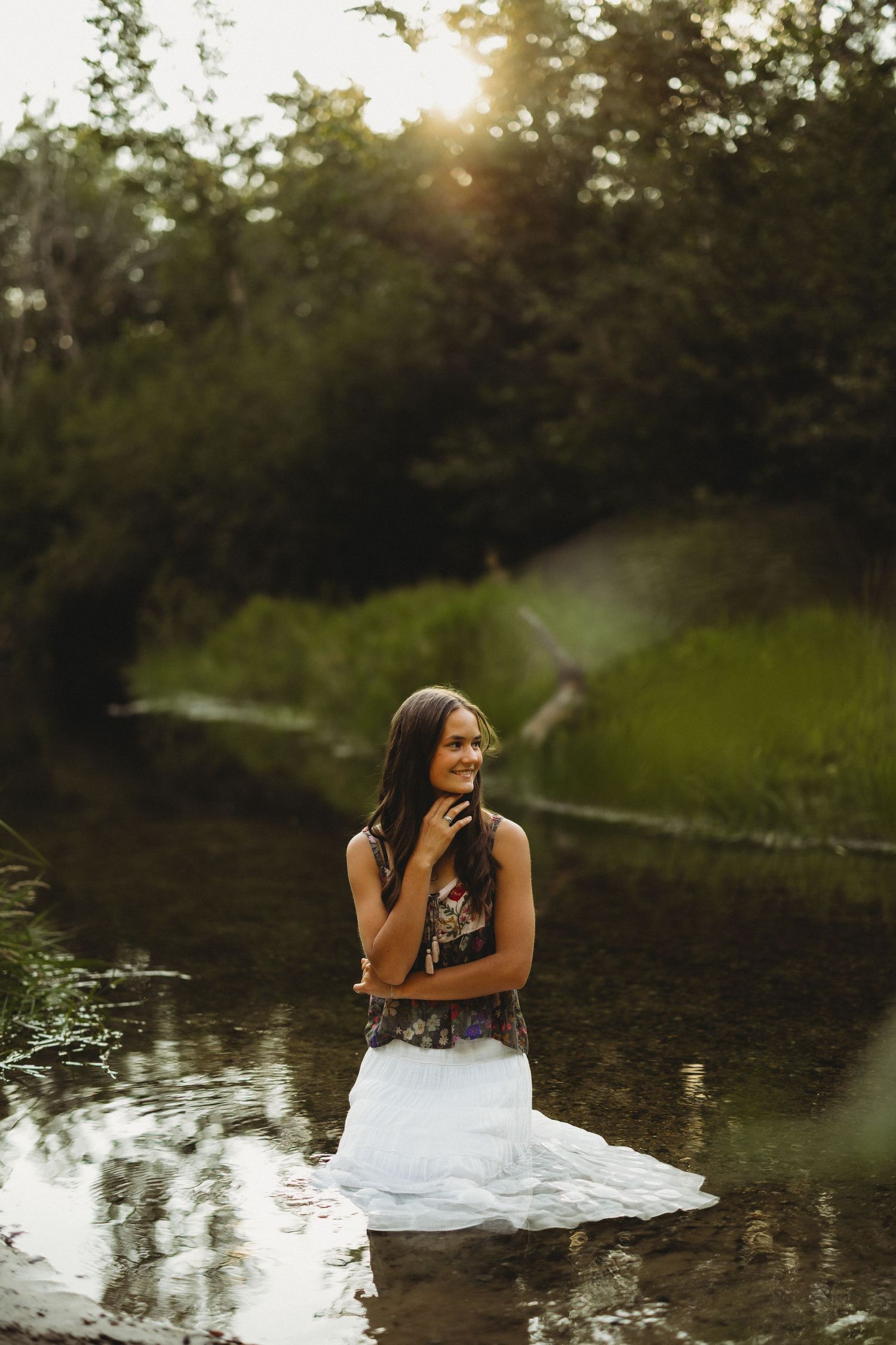 Woman in a white skirt and floral top stands in a shallow stream, smiling softly as golden sunset light filters through green trees.
