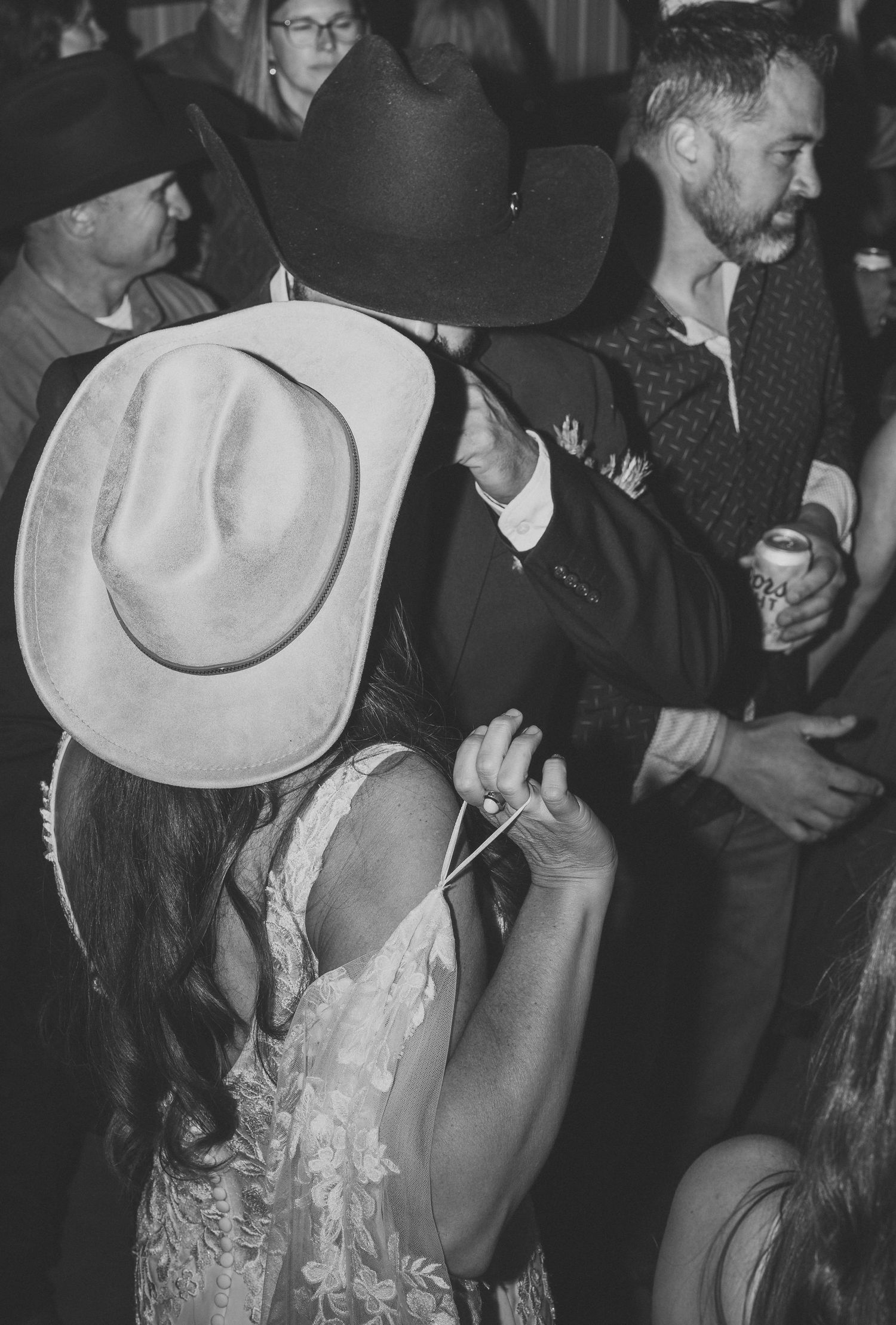 Bride in a lace wedding dress and cowboy hat embraces a groom in a dark cowboy hat on a crowded dance floor at a western wedding.