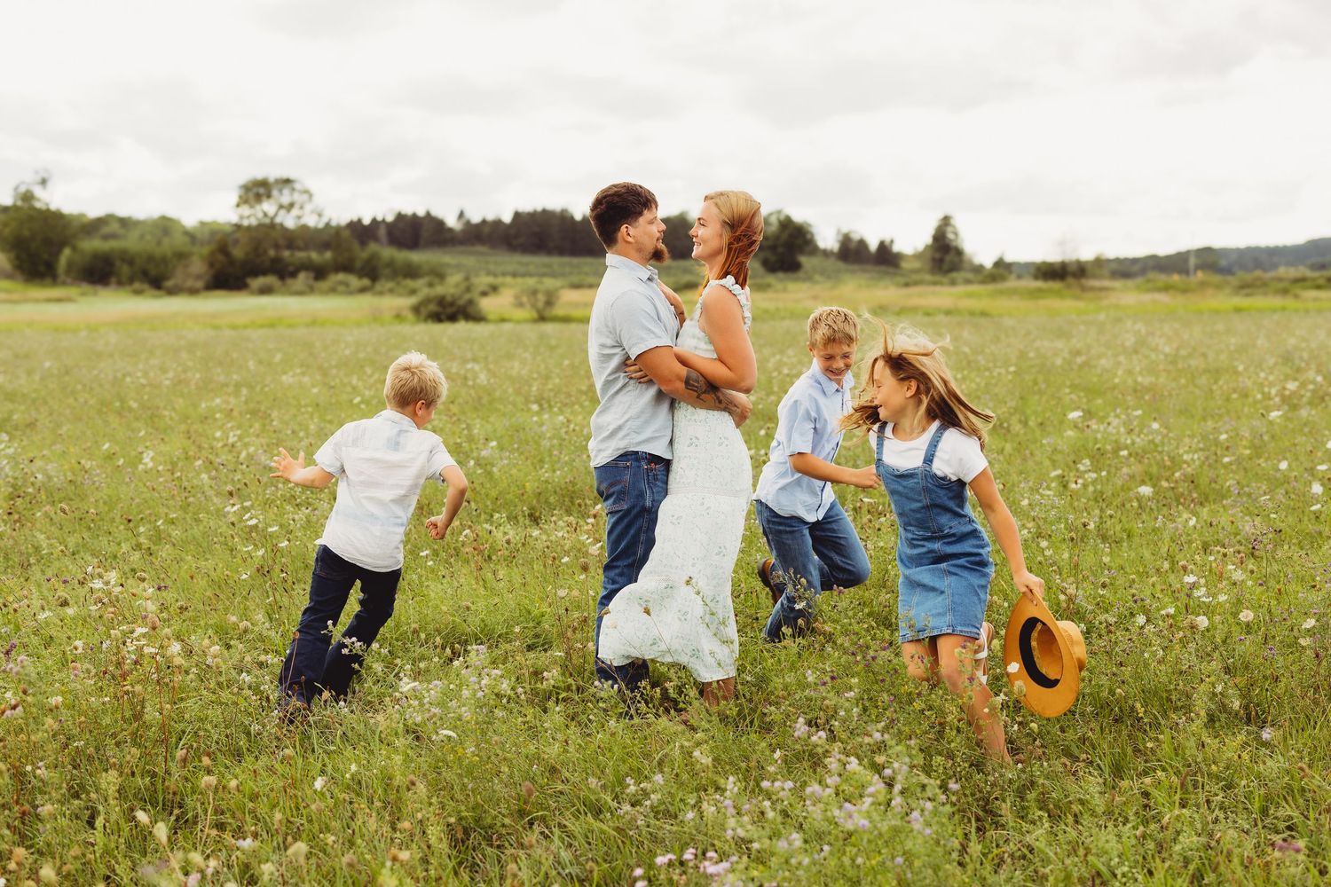 Happy family of five embracing and playing together in a sunny green meadow filled with wildflowers, with children running nearby.